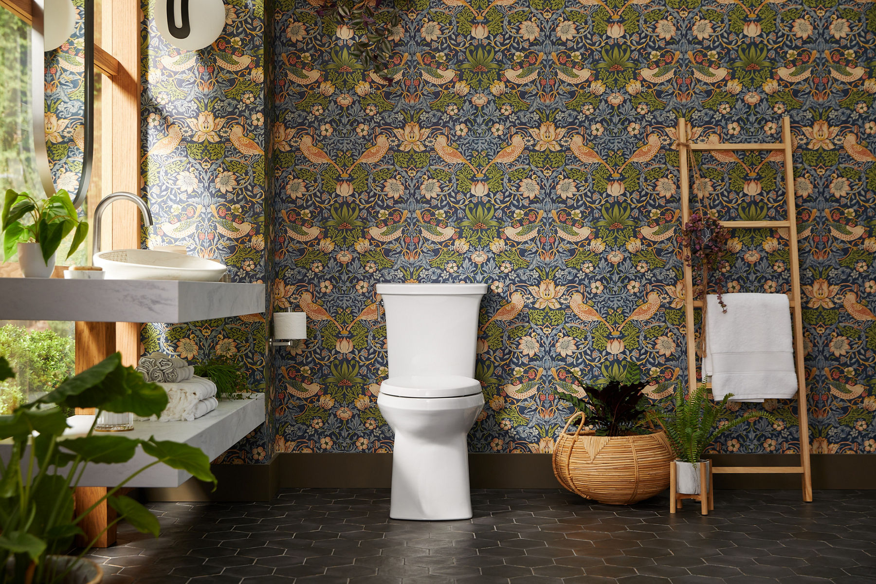 An eclectic bathroom with bird and flower wallpaper, white KOHLER toilet, towel ladder, and lots of potted plants.