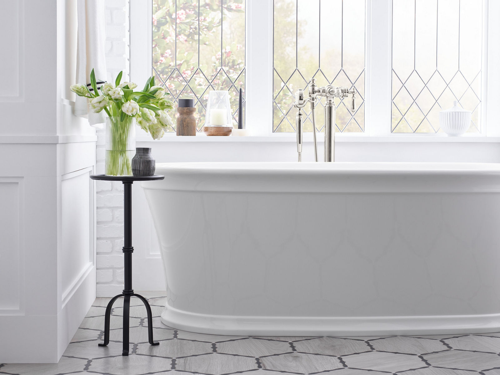 A vase of flowers sits on a candelstick table beside a white KOHLER Memoirs freestanding oval bath.