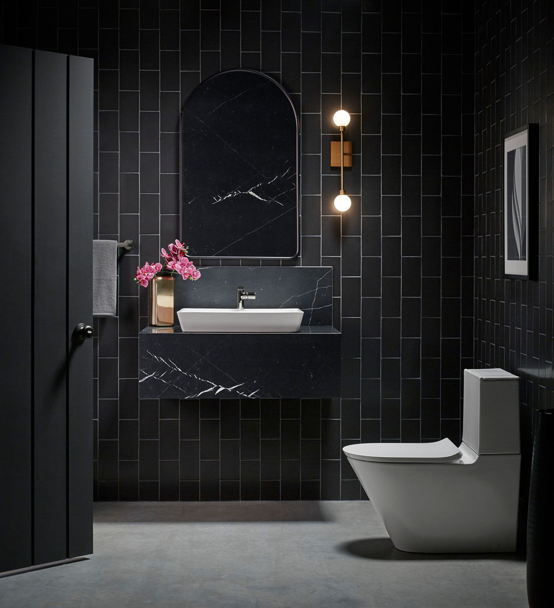 A white Brazn sink and toilet in a modern bathroom with vertically-oriented rectangular black tile with light gray grout, a black quartz wall-hung vanity and dark pink orchids.