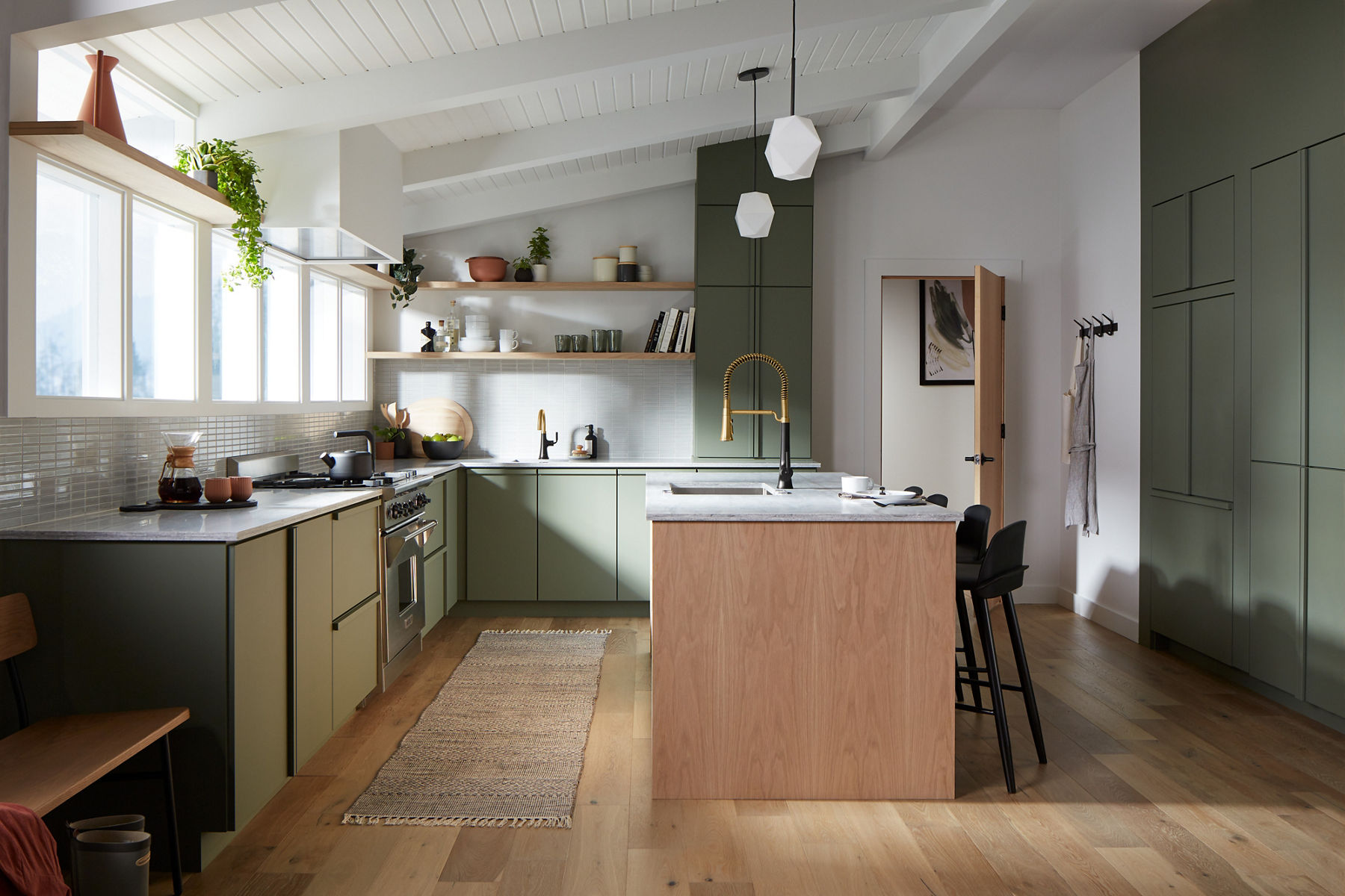 A row of large windows light a kitchen with l-shaped counters, and a large island with sink and KOHLER Tone pull-out faucet in Matte Black with Gold accents.