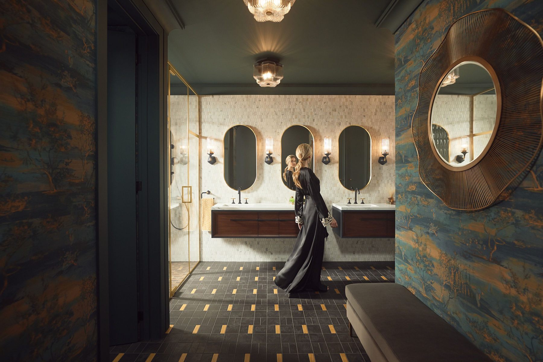 A blonde woman in a long black dress checks her reflection a bathroom with two KOHLER Lodern wall-mounted vanities, Occasion wide spread lever handle faucets, and three Essential capsule mirrors.
