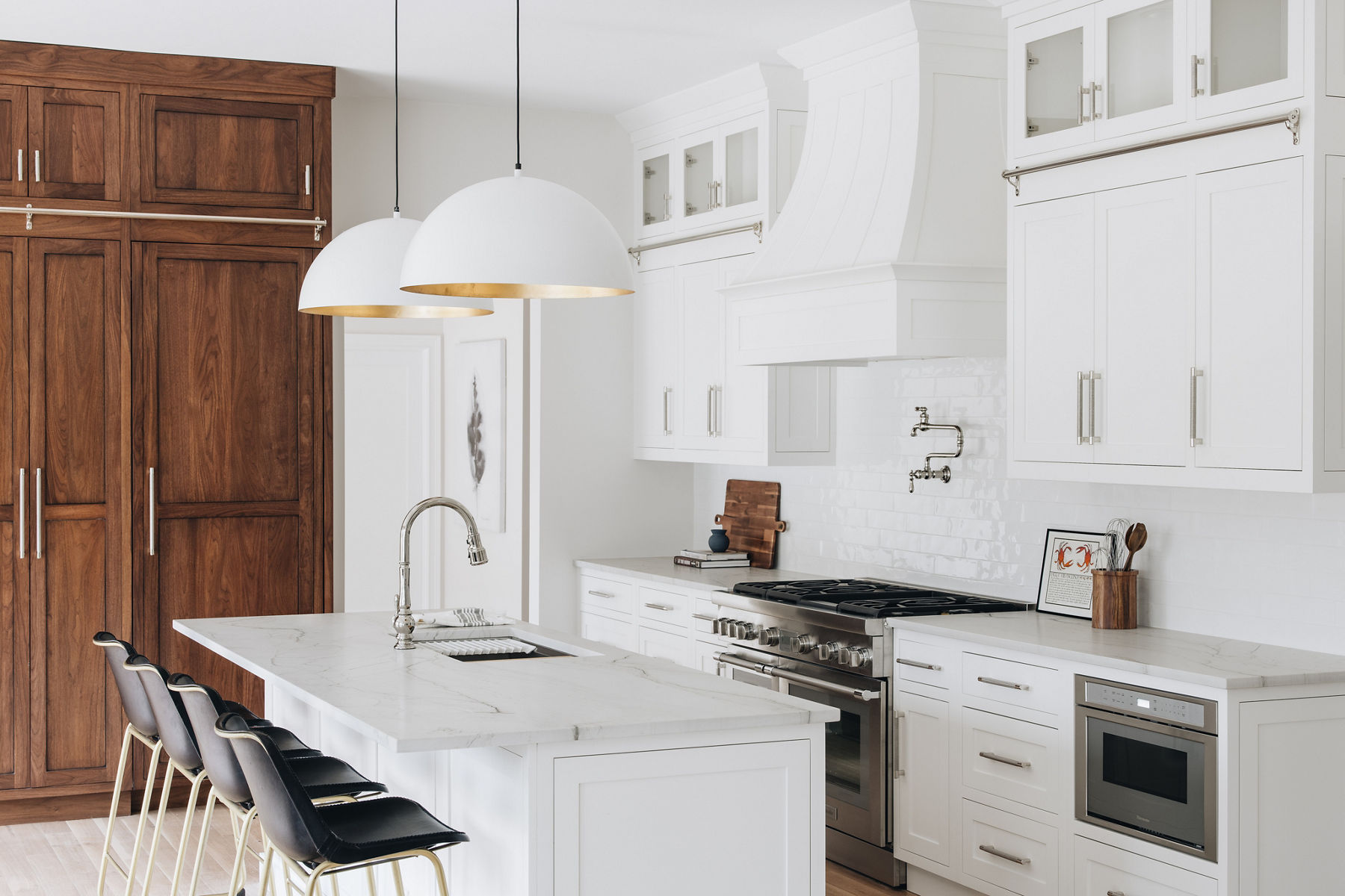 Four black bar stools sit in front of a white island with sink and KOHLER single hole faucet, in a white kitchen with large white range hood and pot filler faucet.