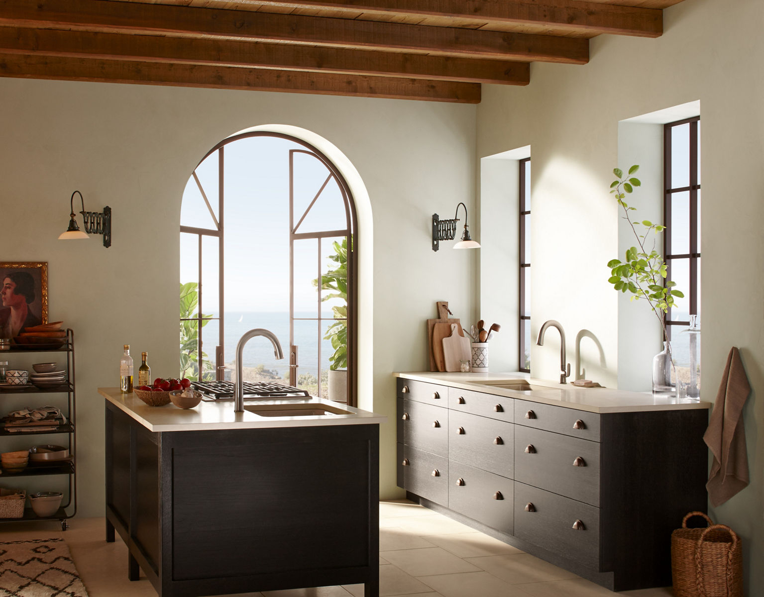 Light streams through a large arched window in a kitchen with ceiling beams, a large island, and a KOHLER Sensate touchless faucet with pull-down spout.