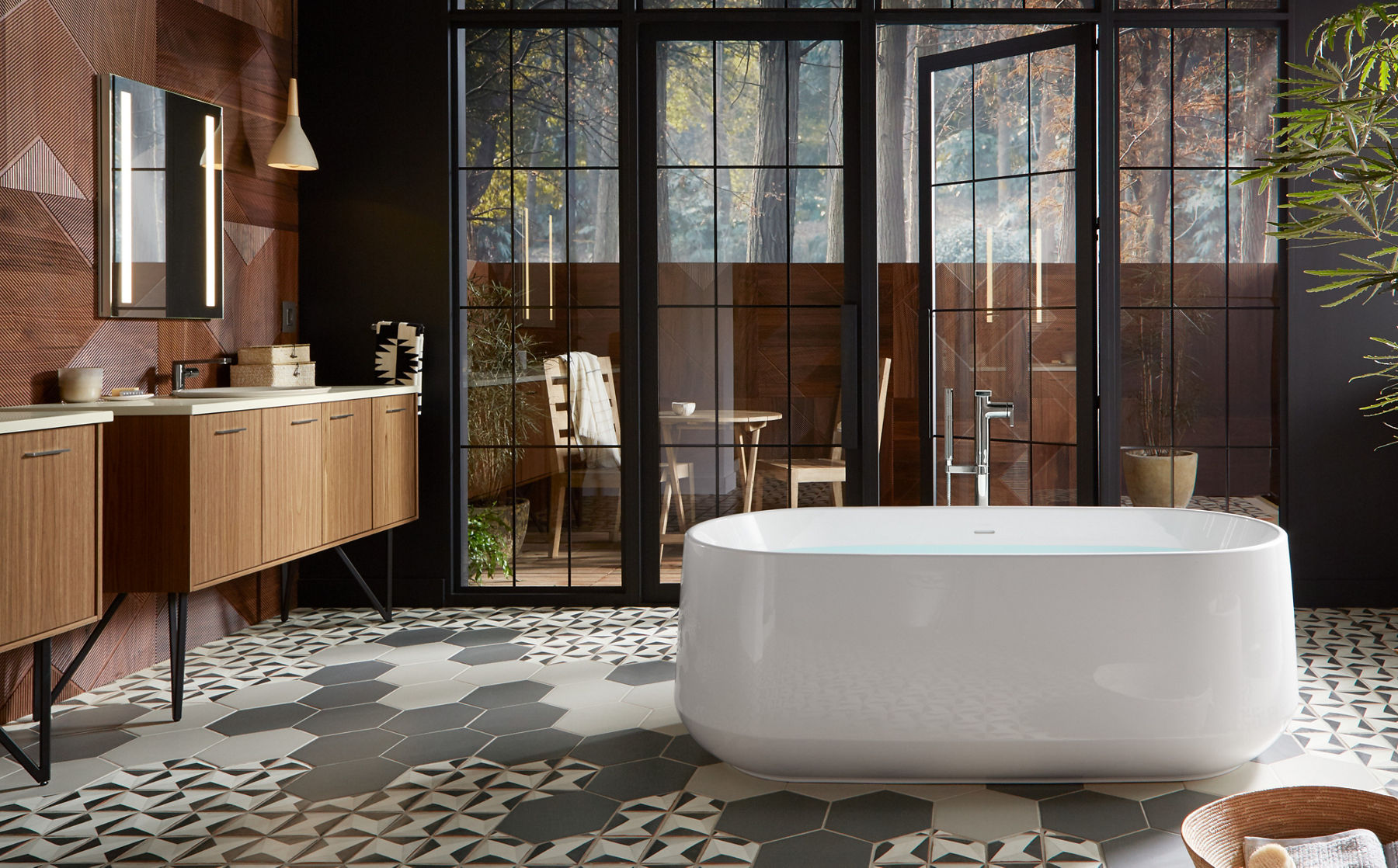A white KOHLER Ceric freestanding bath with Composed floor mount bath filler stands before glass doors leading onto a balcony with a table and chairs in a bathroom with two Jute vanities on hairpin legs.
