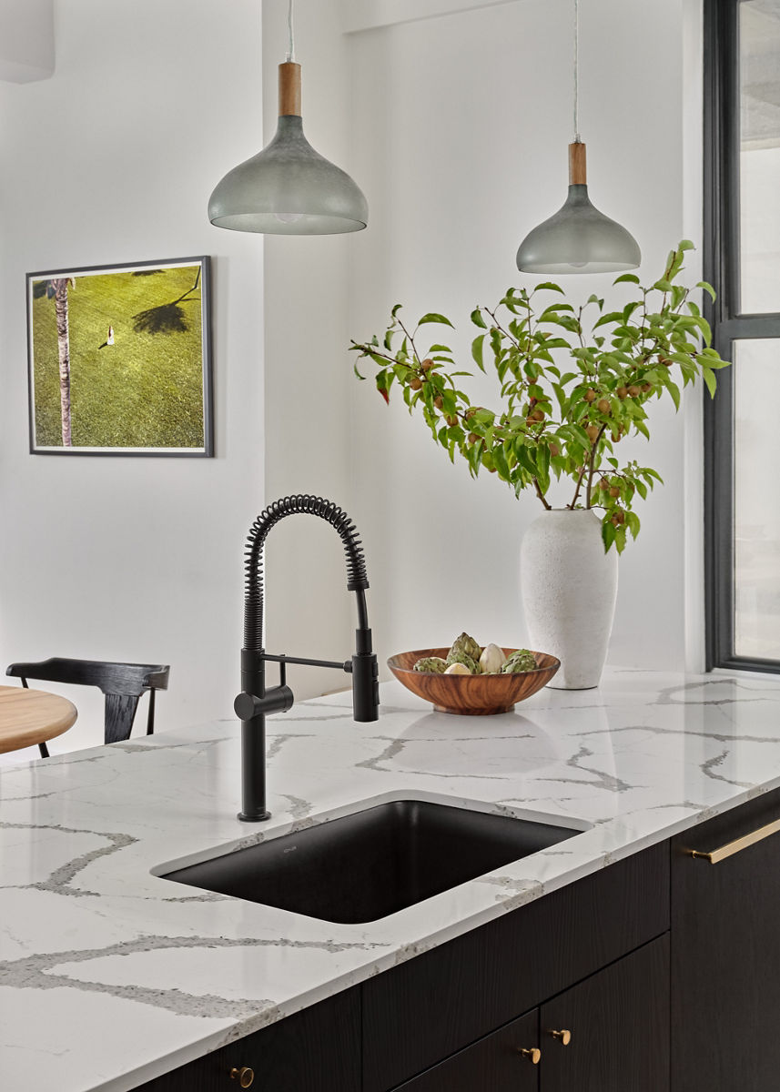 A white vase with green branches stands on a black kitchen island with white and gray marble top, Matte Black KOHLER Crue Semi-Professional faucet, and a black Cairn Neorock undermount sink.