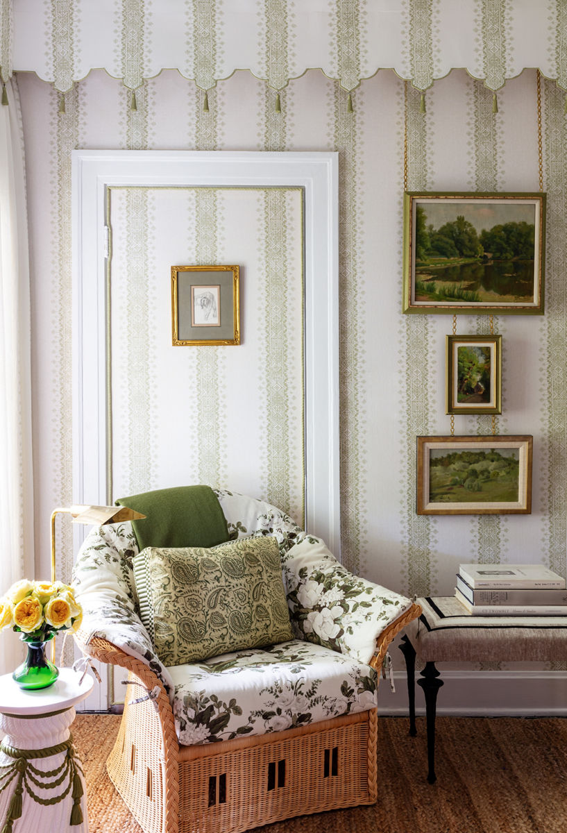 A cozy corner features framed landscape  paintings, green and white striped wallpaper, and a wicker armchair with green and white cushions beside a round pedestal table with a vase of yellow flowers.