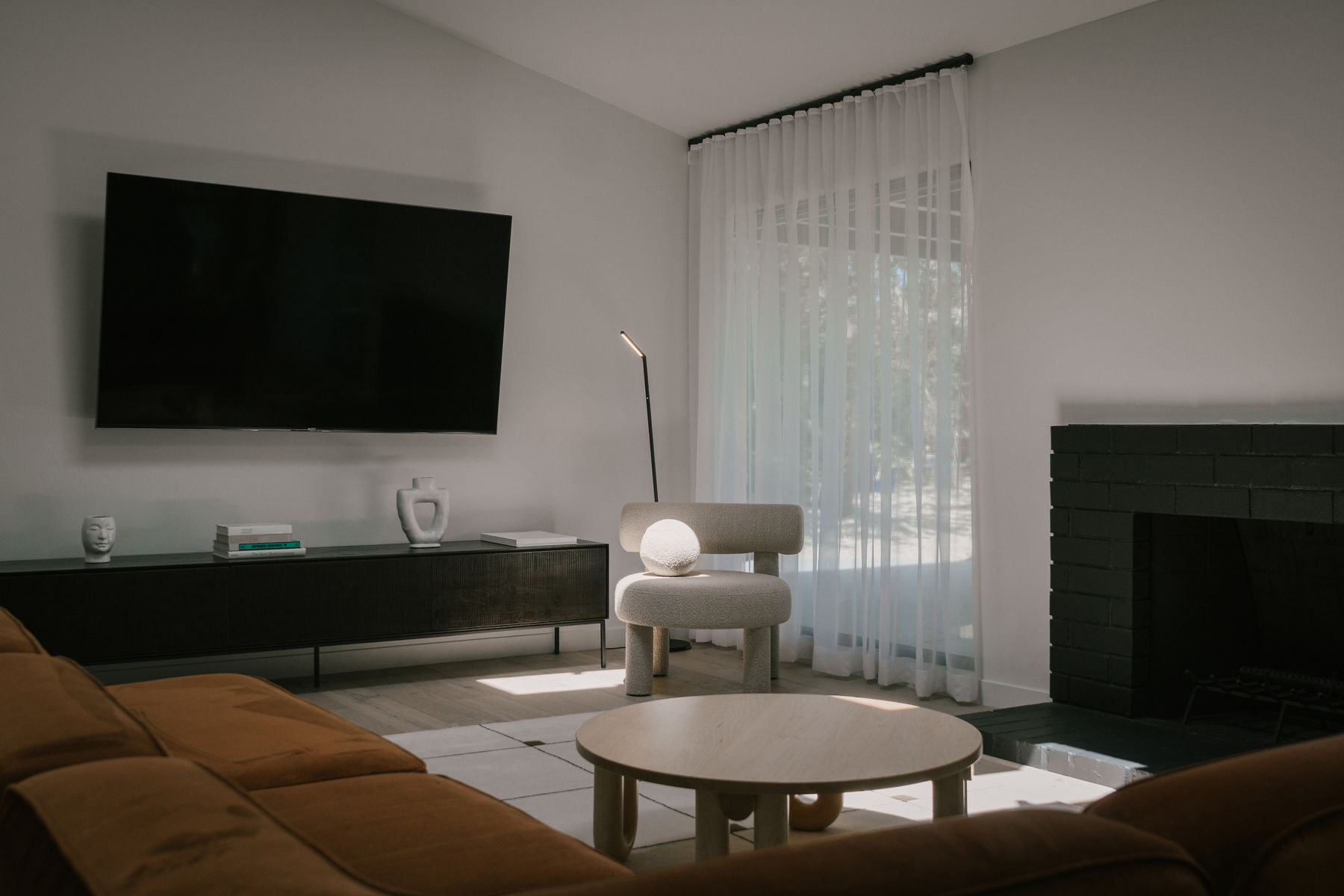 A living room in Japandi style features neutral colors, contrasting textures, a round coffee table, low upholstered chair, rust colored sofa, and black media stand.