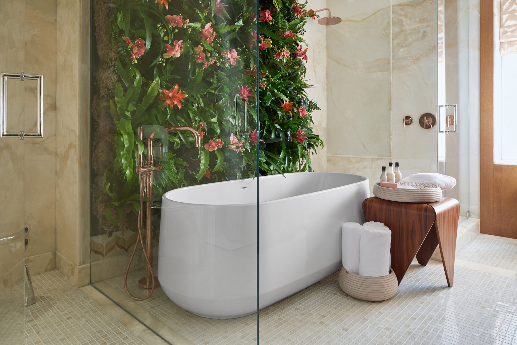 Une salle de bains de couleur crème avec une baignoire autoportante blanche, un mur végétal à gauche et une table en bois avec des accessoires de bain à droite.