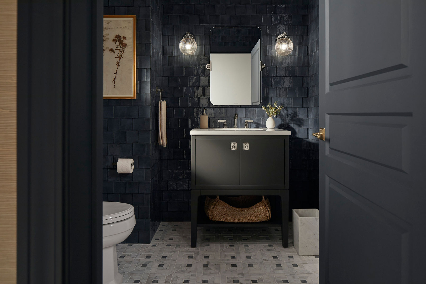 Un meuble-lavabo gris foncé, un robinet chromé, un miroir et des appliques lumineuses conçus par Shea McGee dans une salle de bain avec des carreaux muraux bleu foncé.