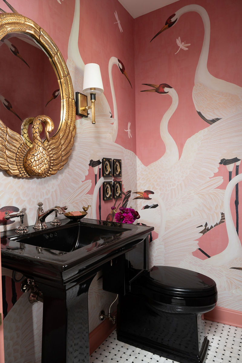 Une salle de bain élégante dotée d’un lavabo et d’une toilette noirs avec un papier peint rose avec motif de cygne, agrémenté d’un vase avec fleurs pourpres sur le comptoir.