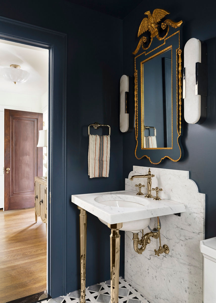 A portrait image of a sophisticated bathroom with dark blue walls, a white marble vanity with brass fixtures, an ornate gold-framed mirror, wall-mounted lights, and a striped hand towel, leading into a hallway with a wooden door and hardwood floors.