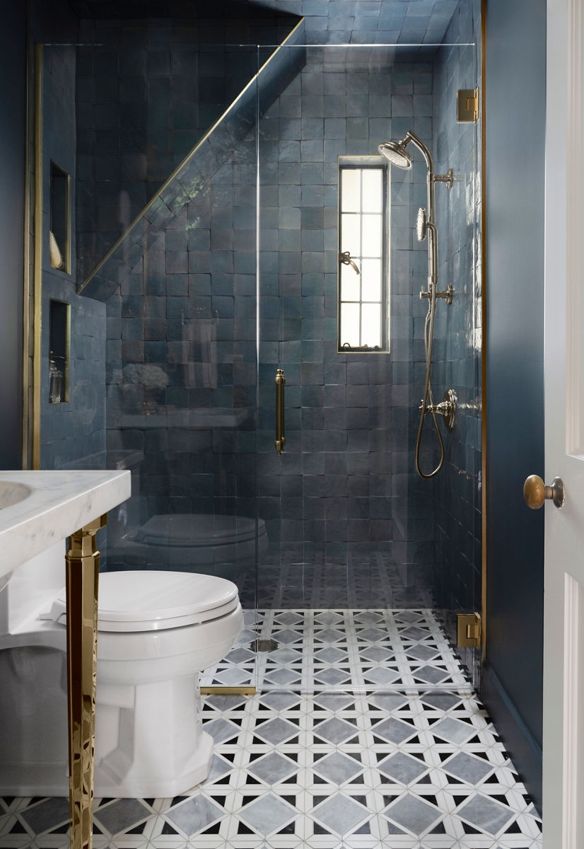A portrait sunlit shot of a modern bathroom featuring a glass-enclosed shower with dark blue tiled walls, brass fixtures, a narrow window and geometric patterned floor tiles, adjacent to a white toilet and marble countertop.