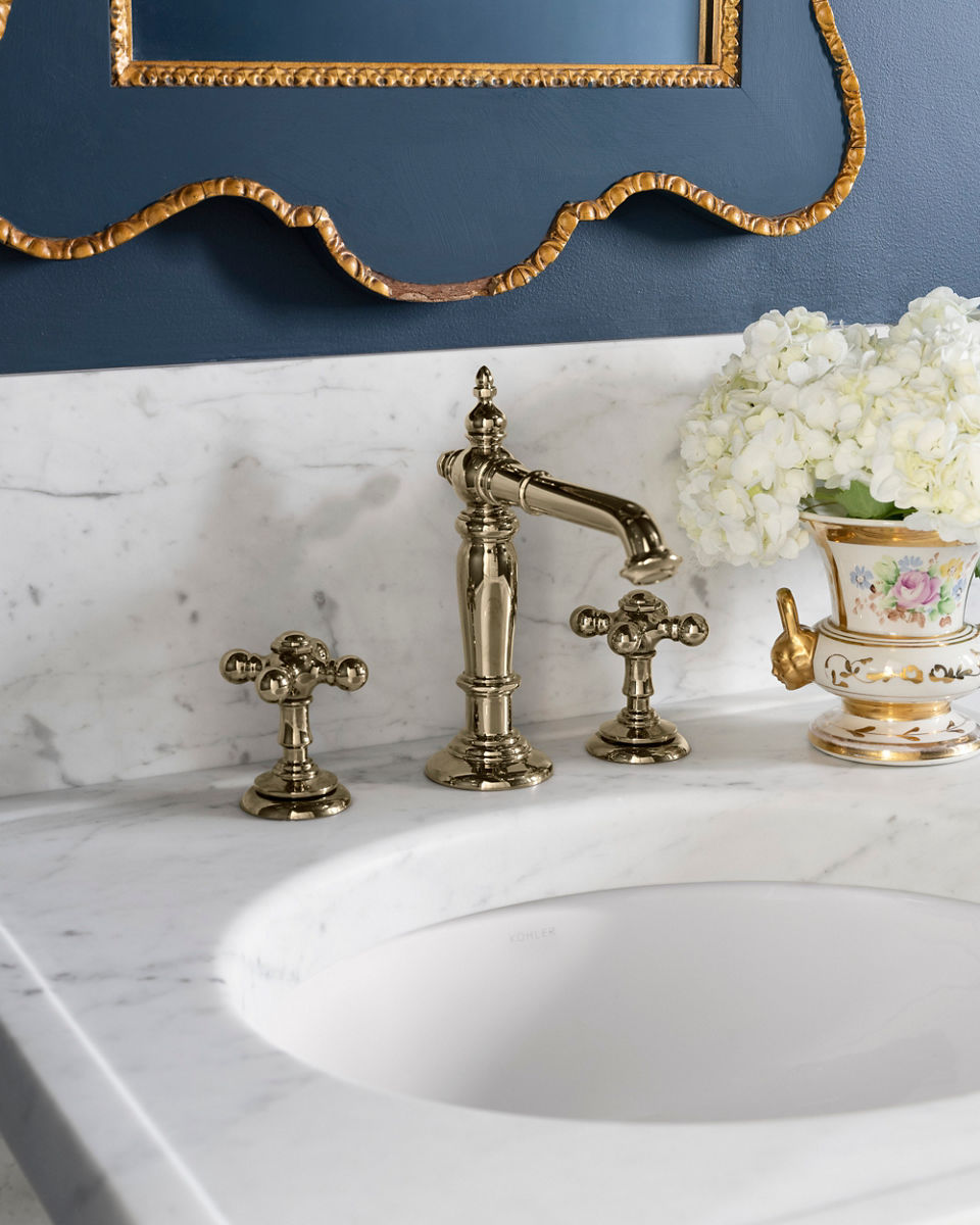 A close-up shot of a luxurious bathroom vanity with a white marble countertop, brass faucet fixtures, a decorative gold-framd mirror, and a floral vase filled with white hydrageas against a dark blue wall.
