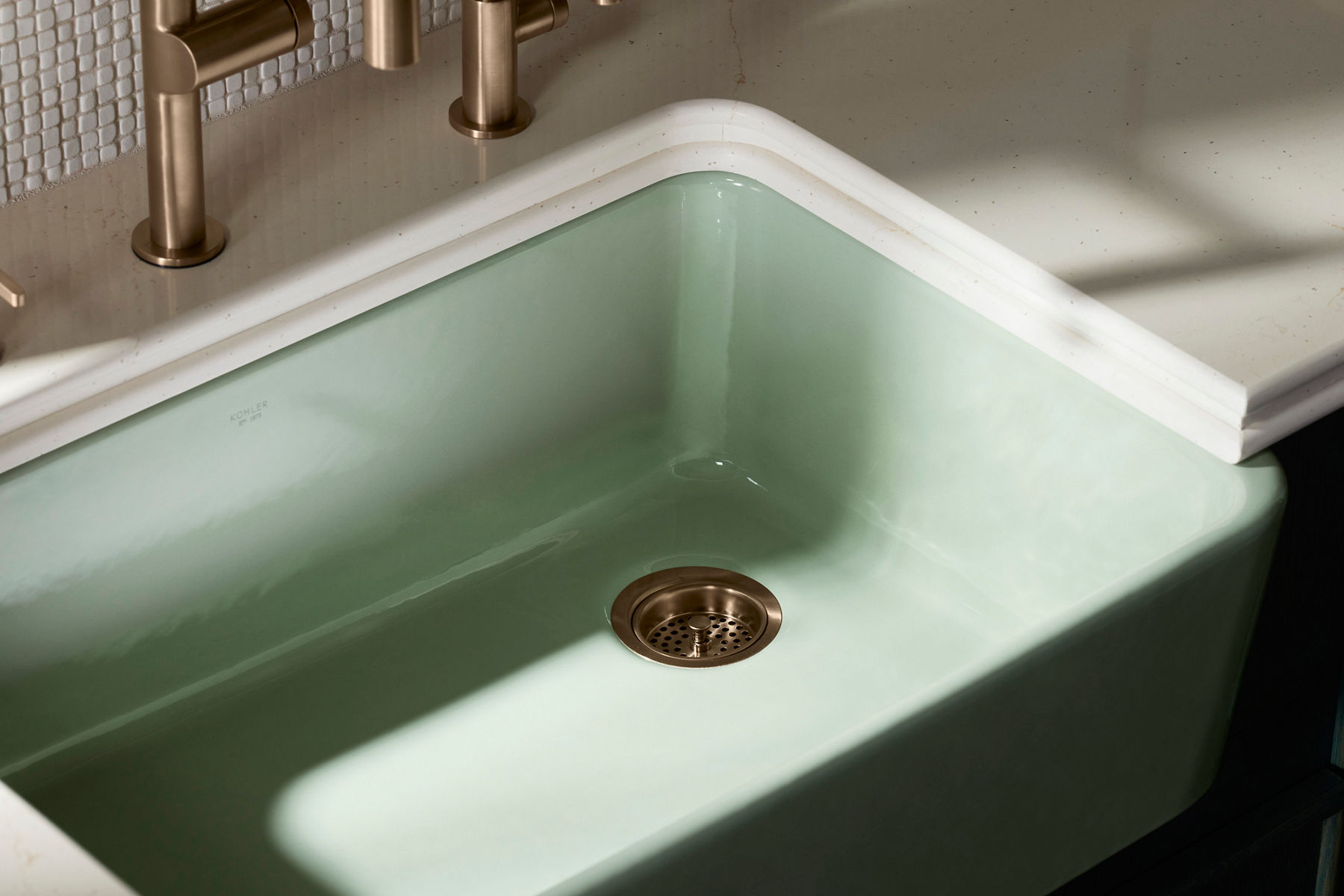 Gros plan sur un drain d’évier doré fixé à un évier de cuisine vert pâle.