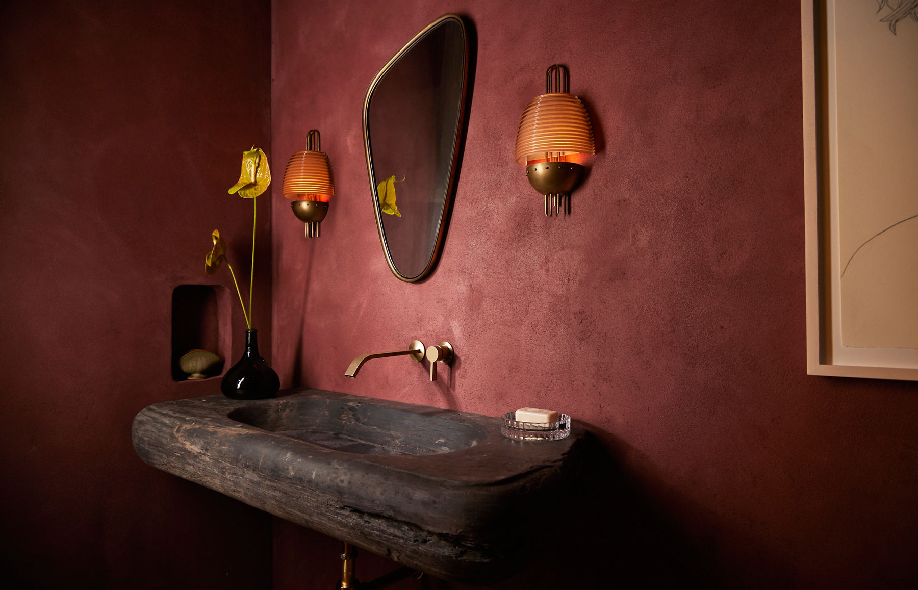 Une image en angle d’un meuble-lavabo en pierre brune avec un robinet doré fixé à un mur marron. Au-dessus du lavabo, on trouve un miroir asymétrique flanqué d’appliques murales orange.