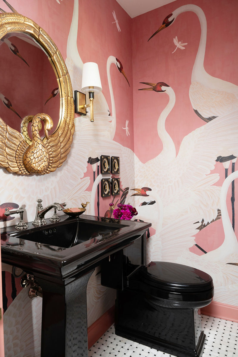 Une salle d’eau élégante dotée d’un lavabo et d’une toilette noirs, d’un miroir en forme de cygne doré et d’un papier peint rose avec motif de cygne.