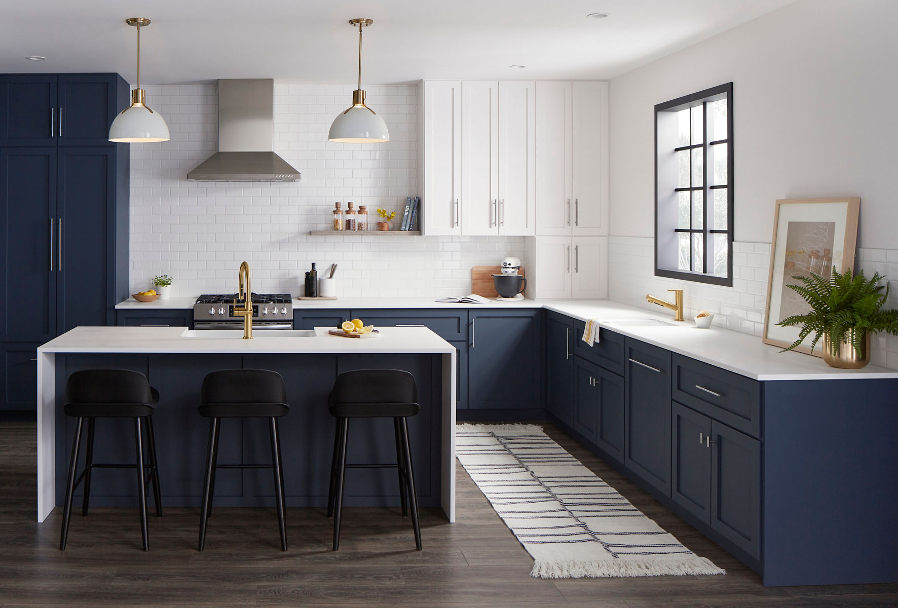 A modern kitchen with navy blue lower cabinets, white upper cabinets, whtie subway tile backsplash, and gold fixtures, featuring a kitchen island with bar stools and various kitchen appliances.