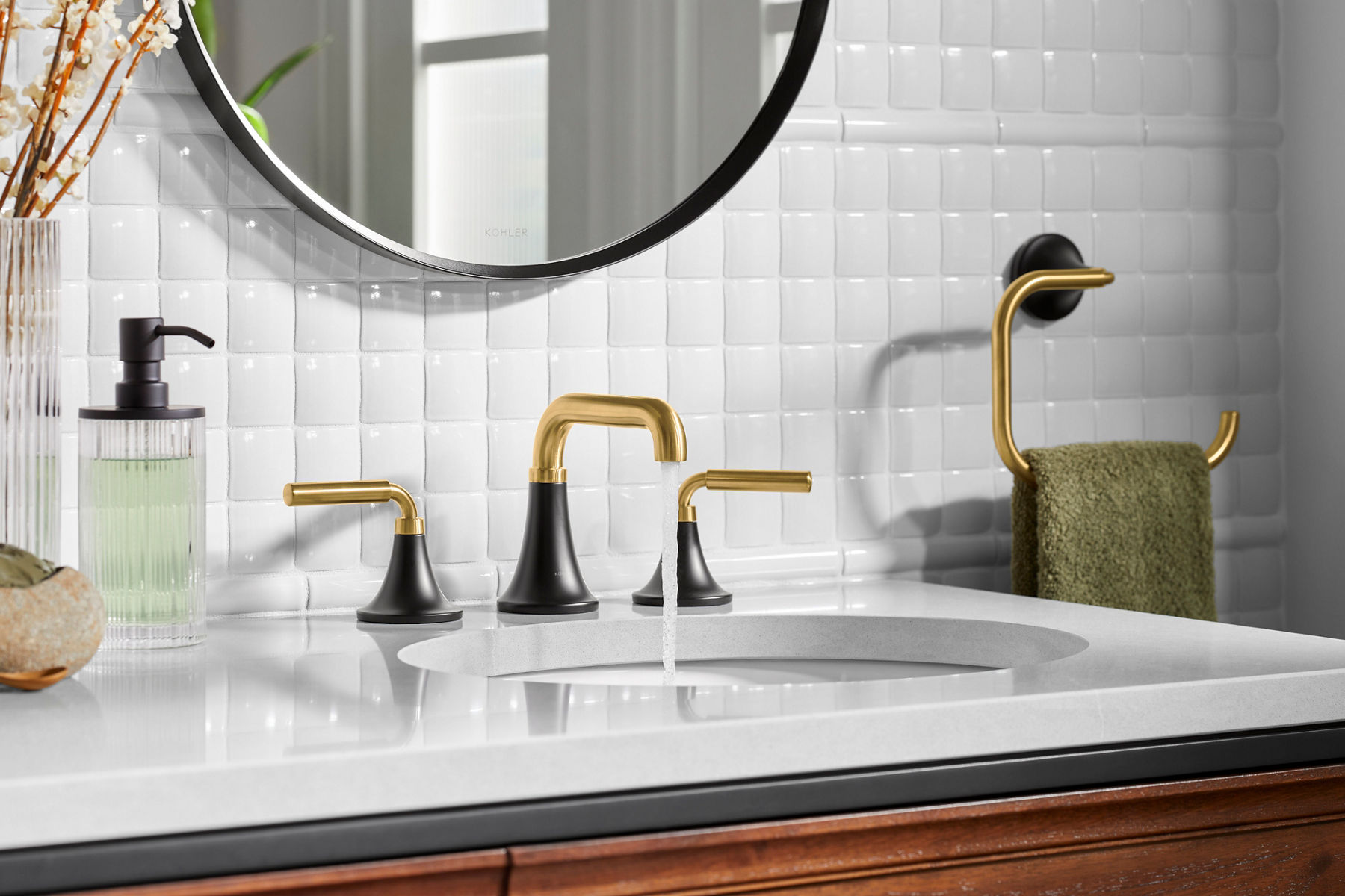 A modern bathroom sink with black and gold faucets, a white tiled backsplash, and a round mirror, featuring a gold towel holder with a green towel.
