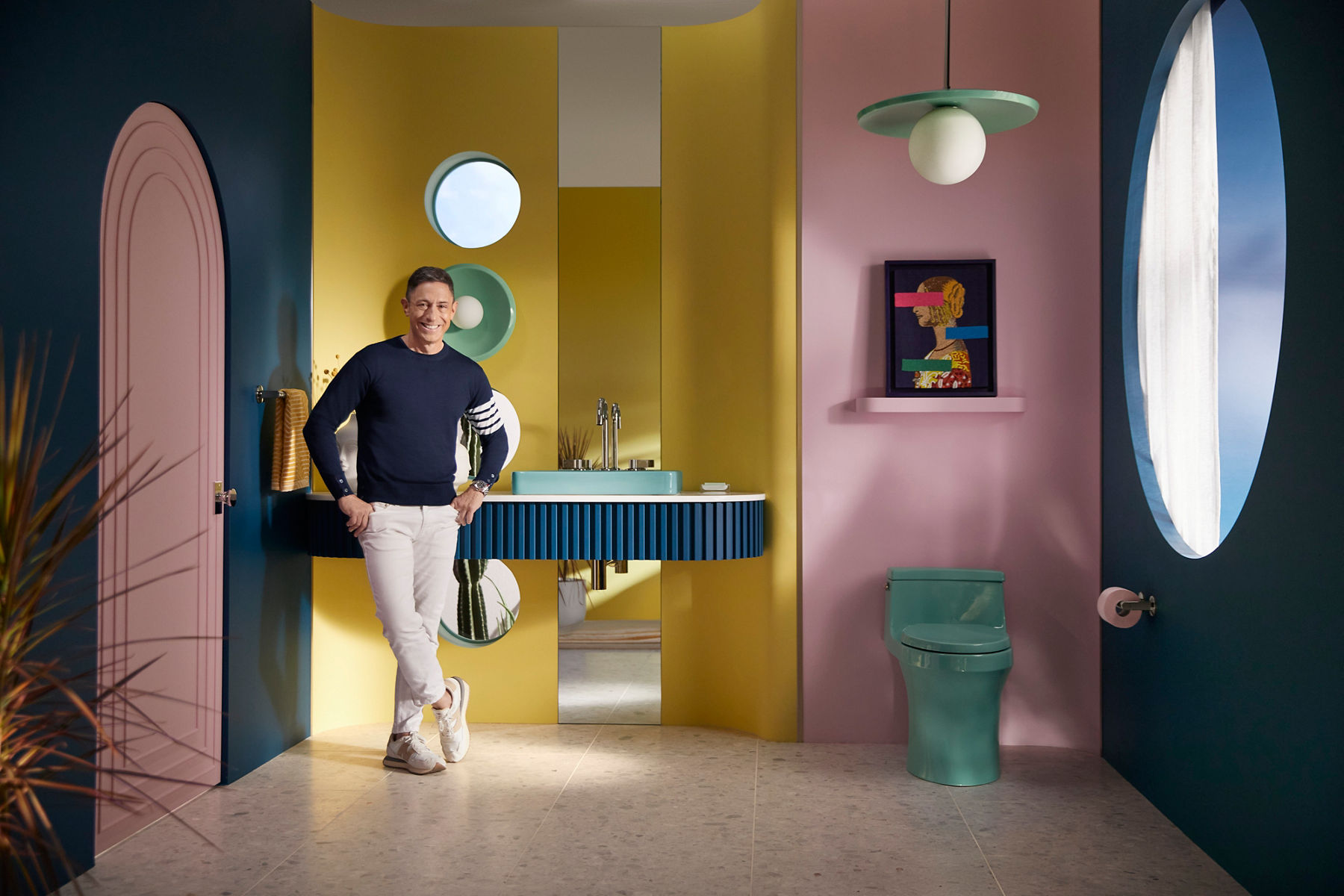 Image du designer et auteur de renom Jonathan Adler debout dans la salle de bain qu’il a conçu avec les nouveaux produits de couleur Heritage de Kohler dans une teinte rétro vert printanier. Il se penche contre le meuble-lavabo avec un lavabo en surface coloré et sourit vers la caméra.