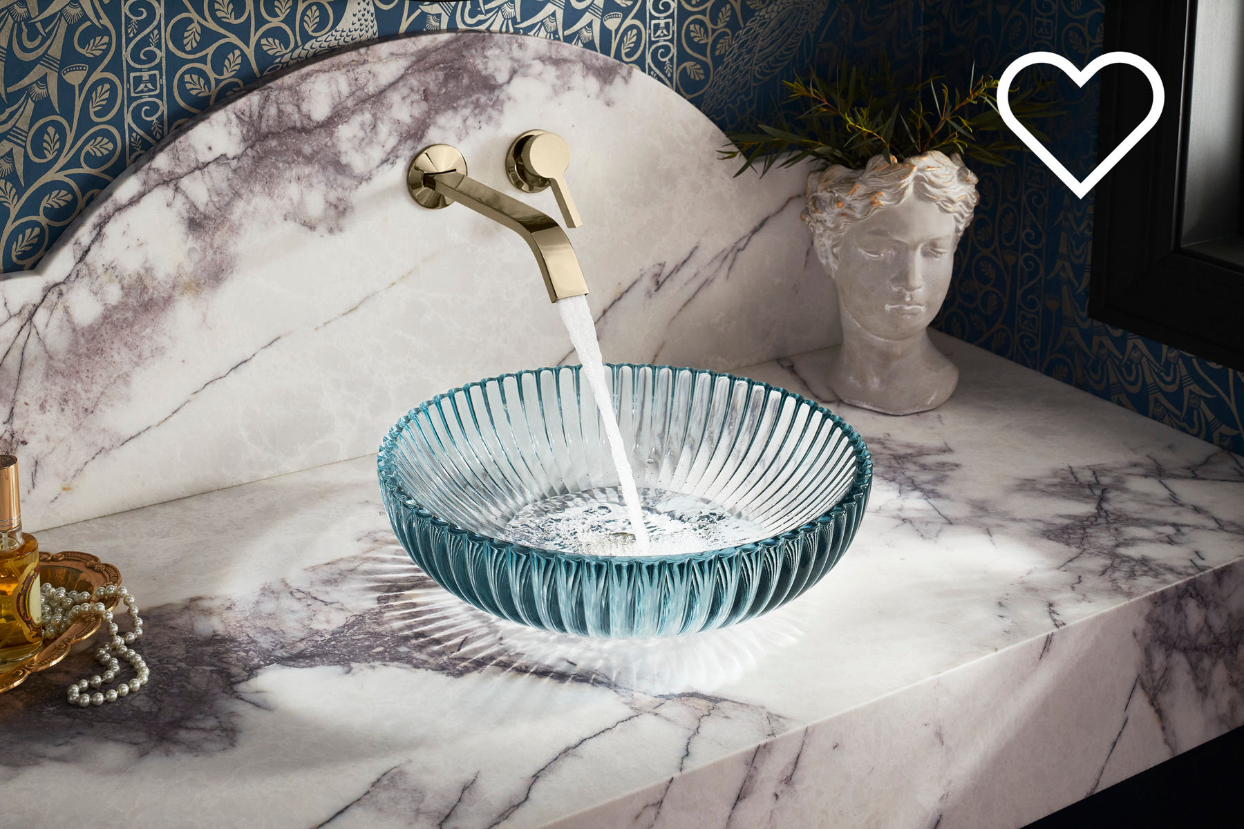 Elegant bathroom vanity with a gold wall-mounted faucet and a blue glass bowl sink set against a marble backsplash and a decorative vase with greenery.