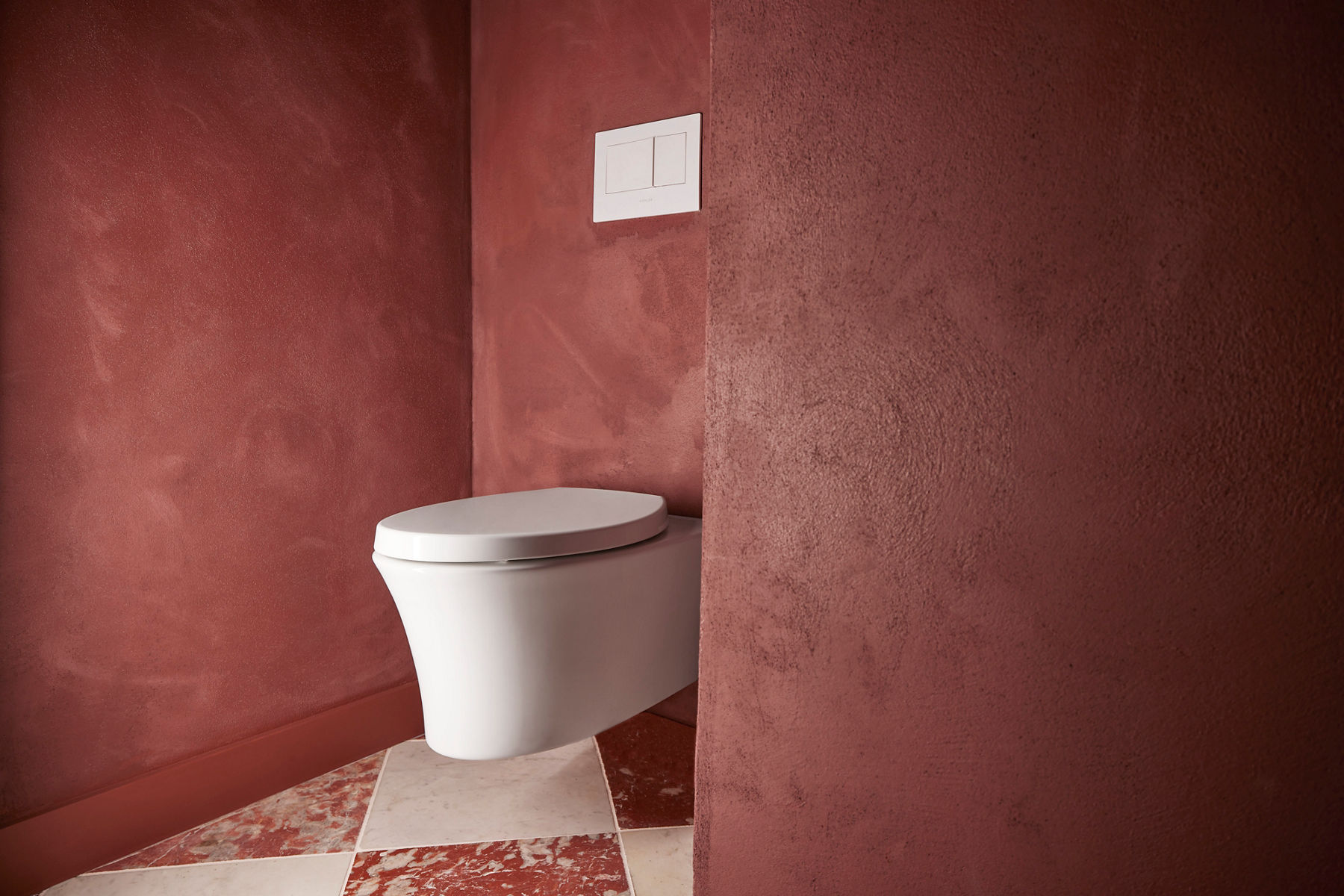 Image en angle d’une salle de bain marron avec une toilette murale blanche. Le siège de toilette est fermé et un levier de chasse blanc est fixé au mur au-dessus de la toilette.