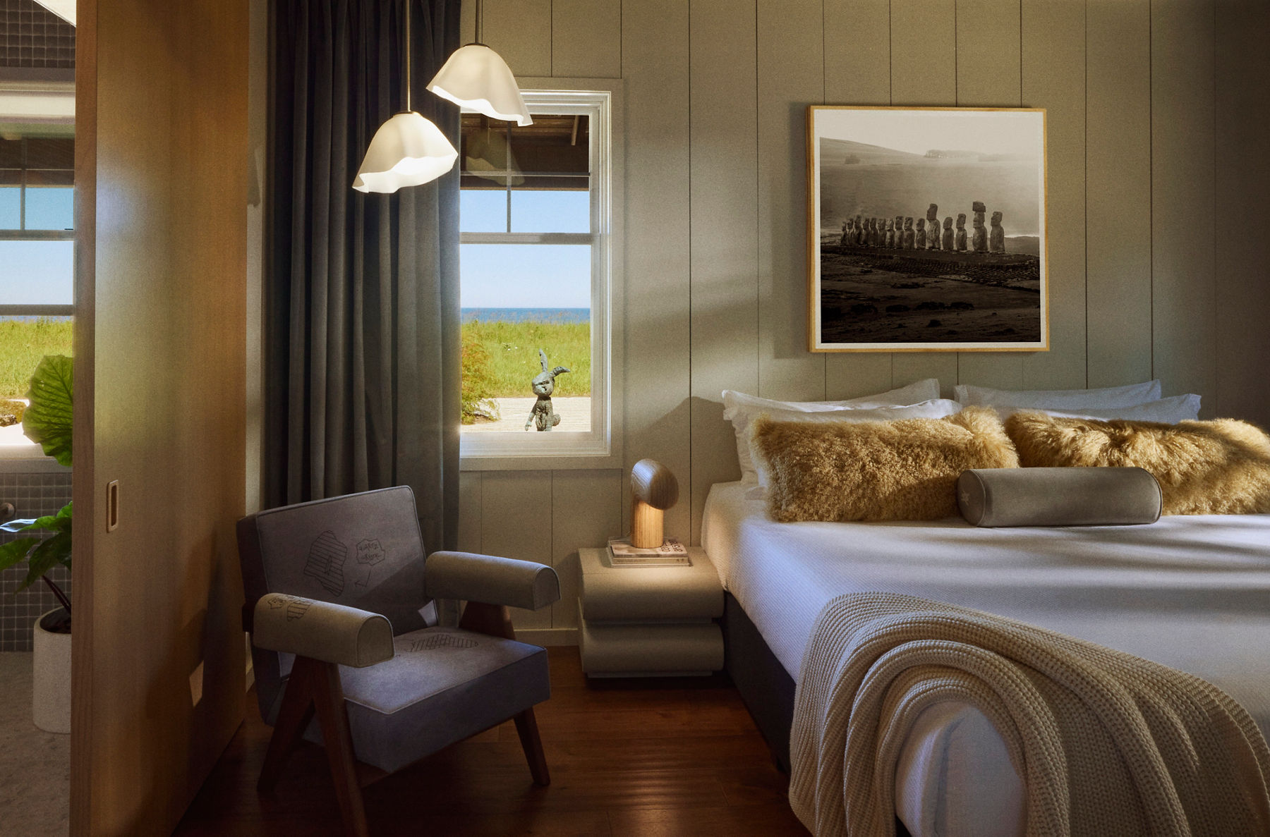 Cozy bedroom interior with a neutral color palette featuring a bed with textured pillows, a side table with a sculptural lamp, a unique chair, and a large framed photograph on the wall. A window reveals an outdoor view of greenery and the ocean.