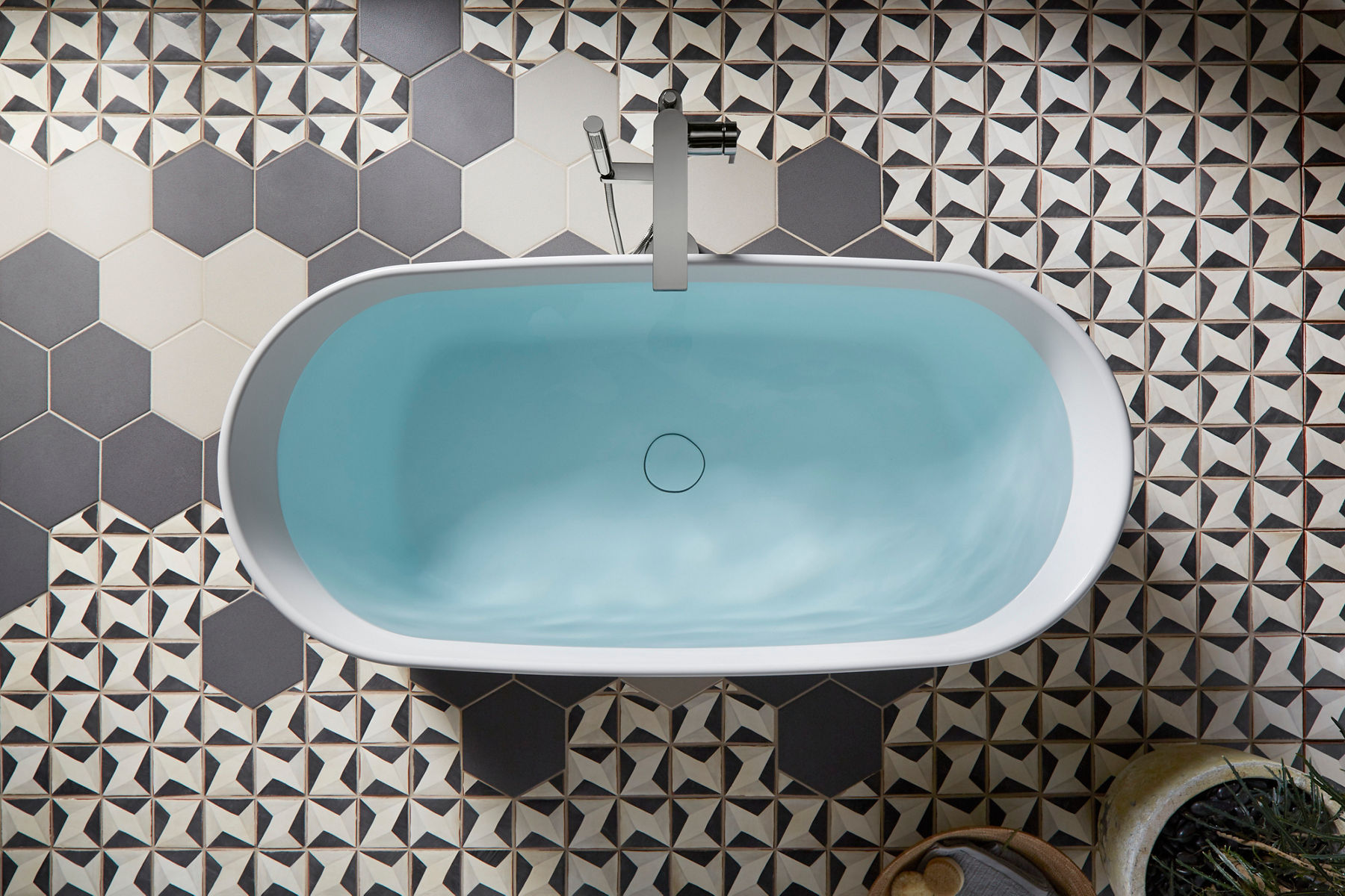 Vue en plongée d’une baignoire autoportante blanche remplie d’eau sur un plancher en carreaux géométriques blanc et gris composés de différents motifs.