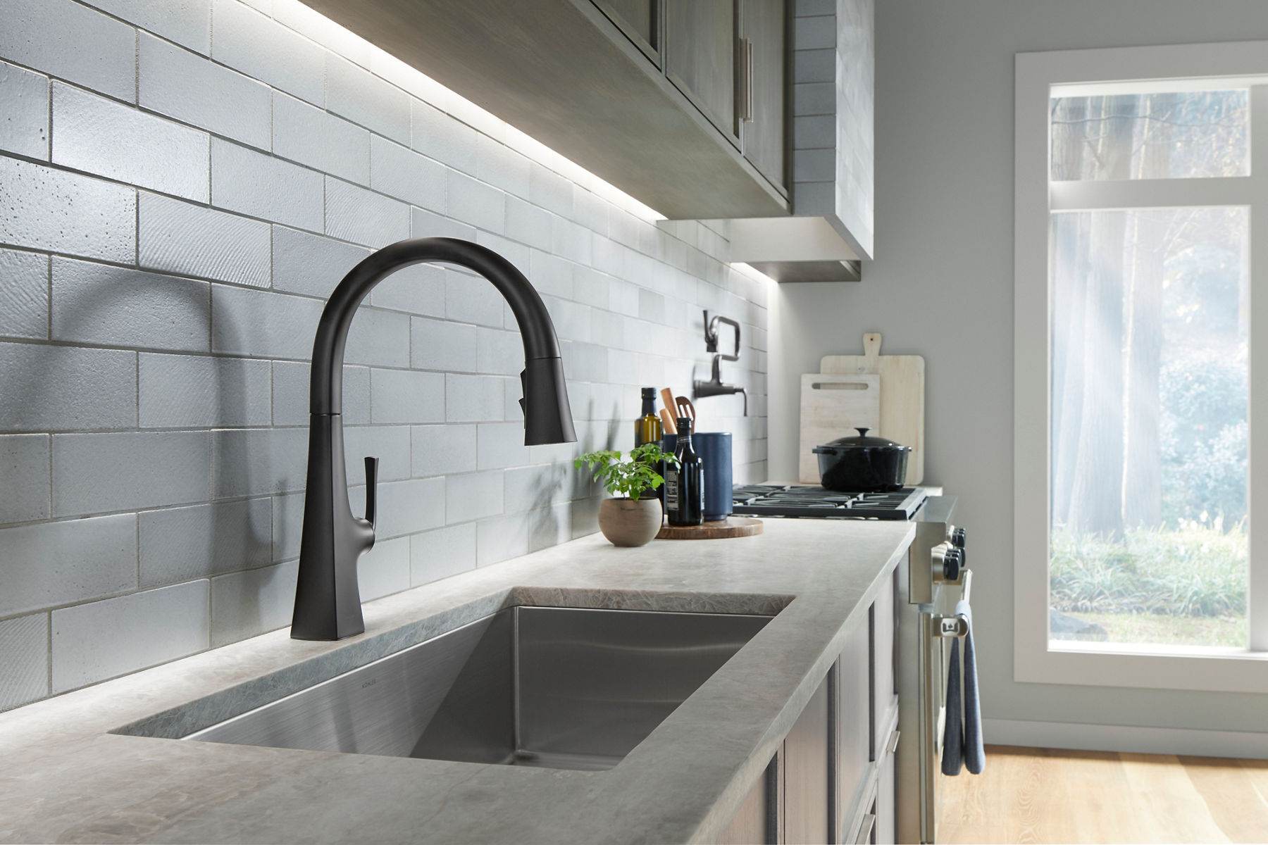 A black faucet with a square base that transitions into a round spout on a gray counter top with a stainless steel sink.