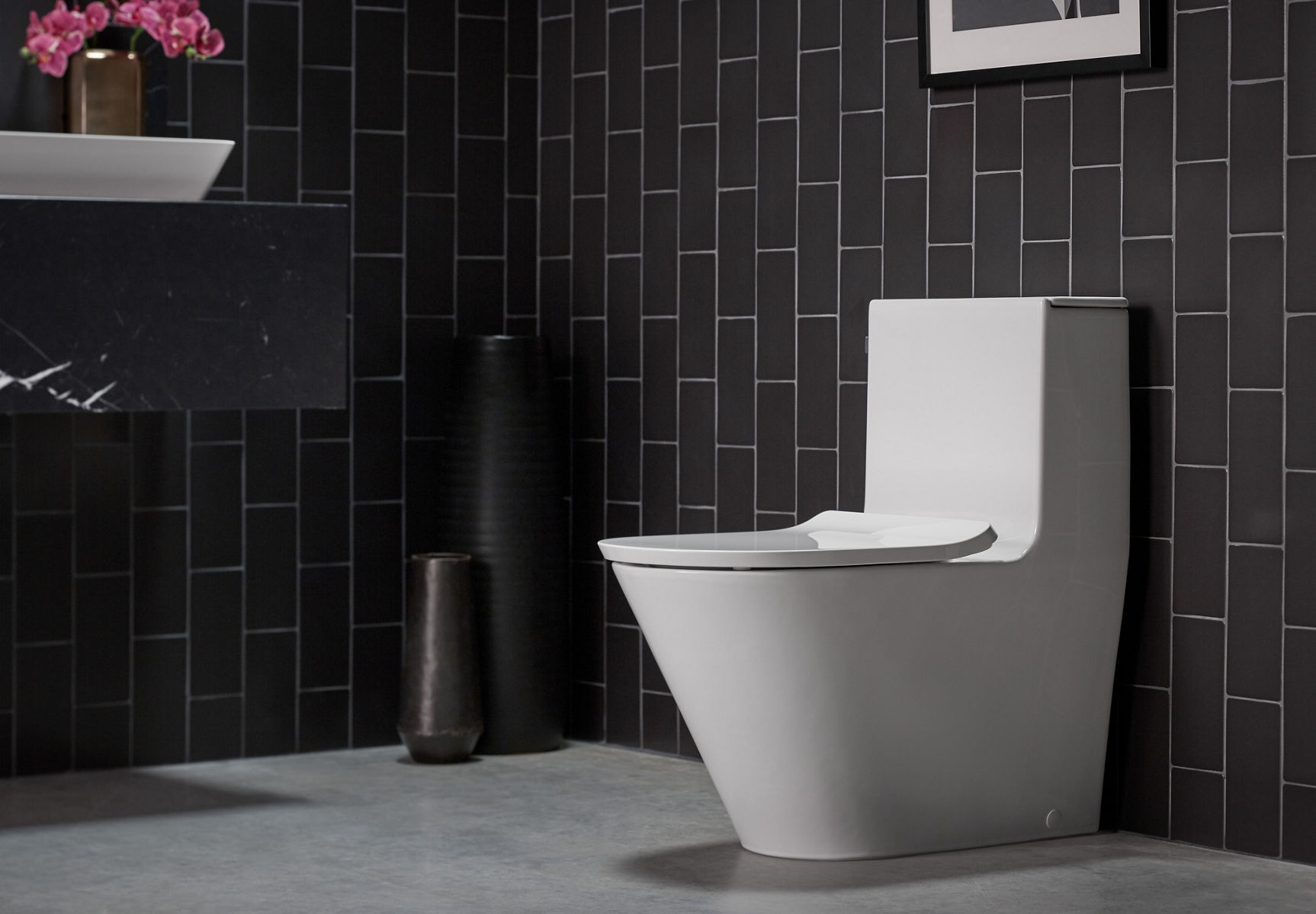 Image of a modern, minimalist bathroom designed in mostly black, white and grey. A white Kohler Brazn one-piece toilet is featured in the middle of the image, against a grey tile flooring and black vertical wall tiles. In the corner of the image, you see the corner of a white Brazn vessel sink on top of a thick marble slab vanity counter top.
