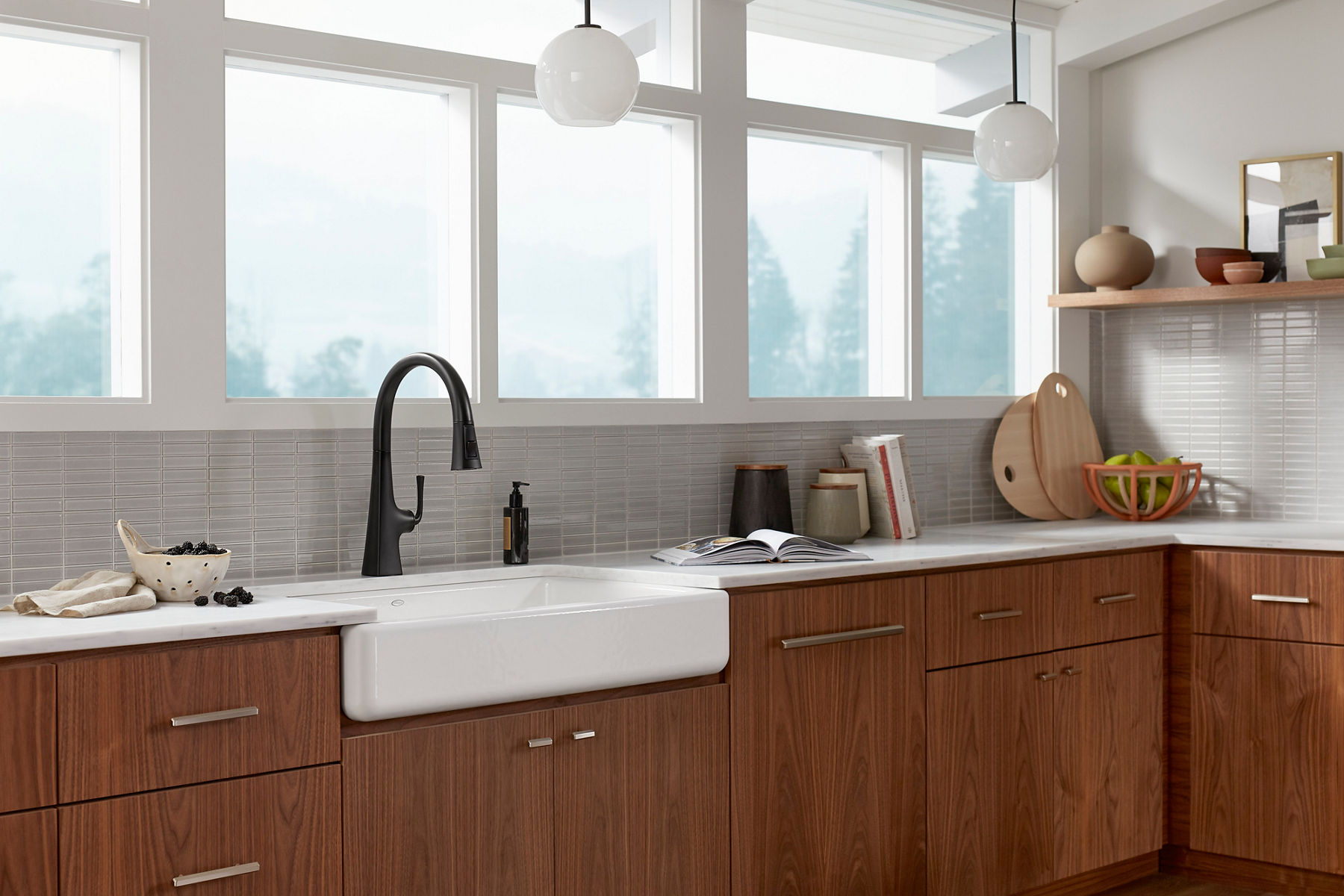  Wide image of an updated kitchen space with natural wood lower cabinets, cream-colored wall tile, and a white farmhouse, apron-front sink as the focal point in the center of the kitchen. A matte black kitchen faucet sits above the sink and in front of a wall of windows, allowing sunlight to fill the room.