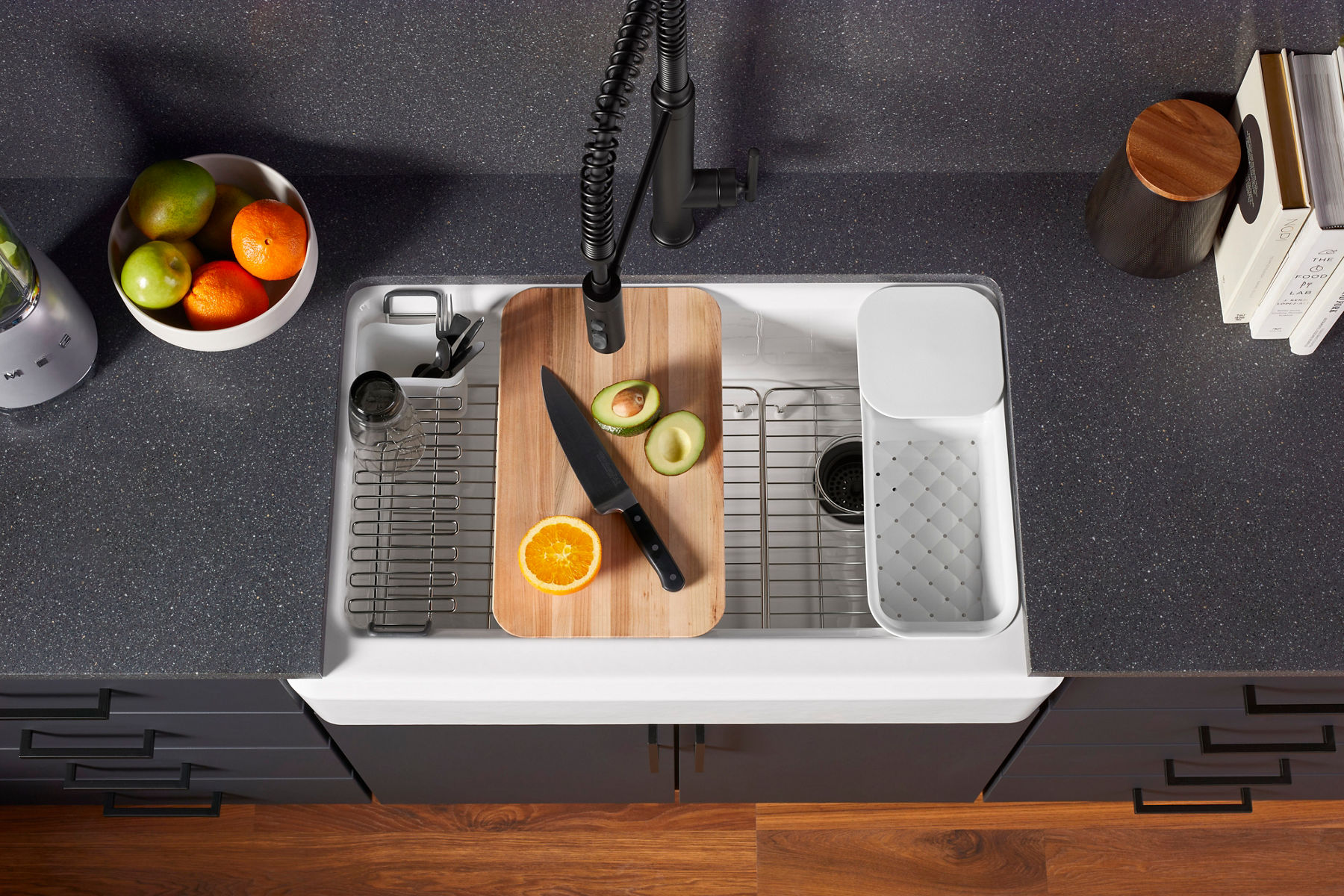 A speckled gray kitchen counter with a bowl of fruit next to a white undermount workstation sink with built-in cutting board, strainer, and other accessories.
