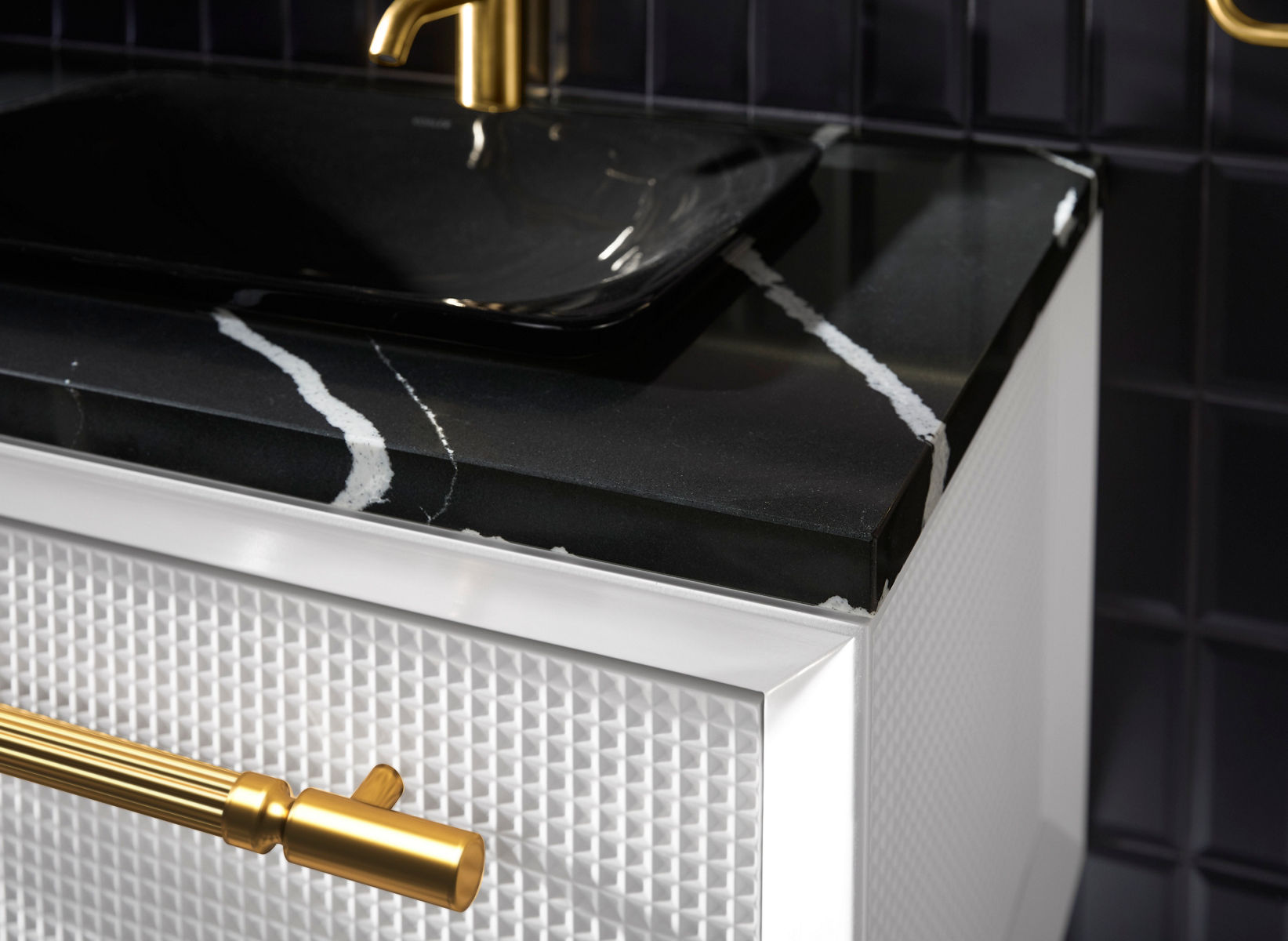 Close-up image of a wall-mounted  Kohler bathroom vanity, showing the detailed texture of the white vanity cabinet, brass drawer pull, black with white veined marble top, black vessel sink and brass single-handle faucet.