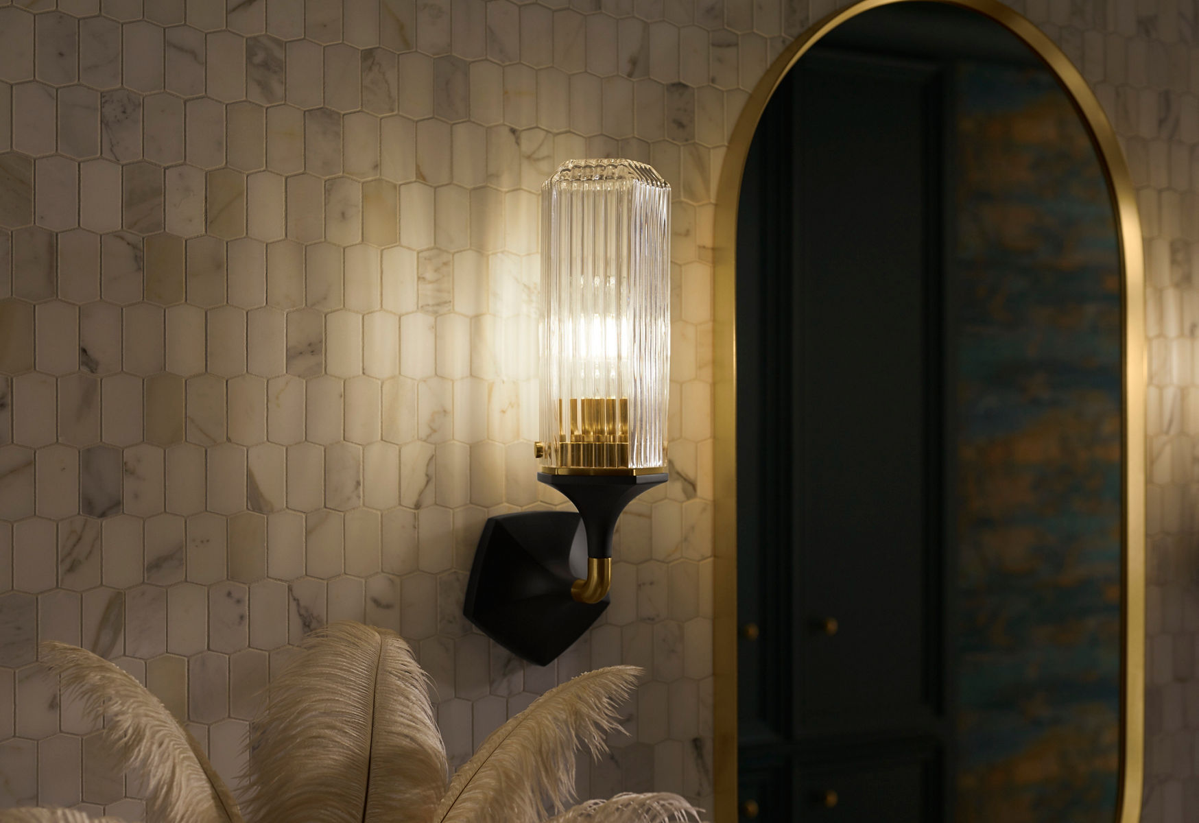 Close-up image of a Kohler Occasion two-tone wall scone in matte black and vibrant brass with a channel patterned glass around the light. Next to the light is an oval Kohler mirror with brass edge. Both are hanging on a small and textured tiled bathroom wall.