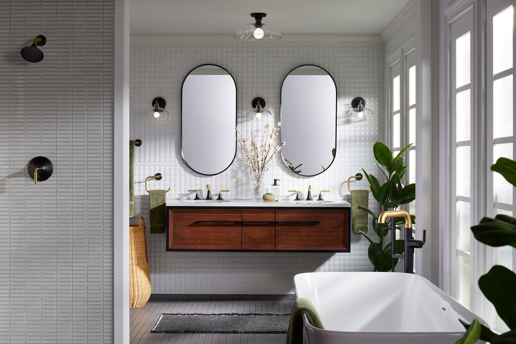 Wide image of a full bathroom suite featuring a double vanity in dark stained wood hung against the wall with a white countertop and white textured wall tile. Matching oval mirrors are hung above the matching Kohler Occasion two-toned faucets in matte black and brass. Three Kohler wall sconces are placed alongside and in between the two mirrors. In the foreground of the image, you can see a large bathtub with a matching Occasion tub filler and a shower with Occasion shower head and control handle, all in two-tone matte black and brass.