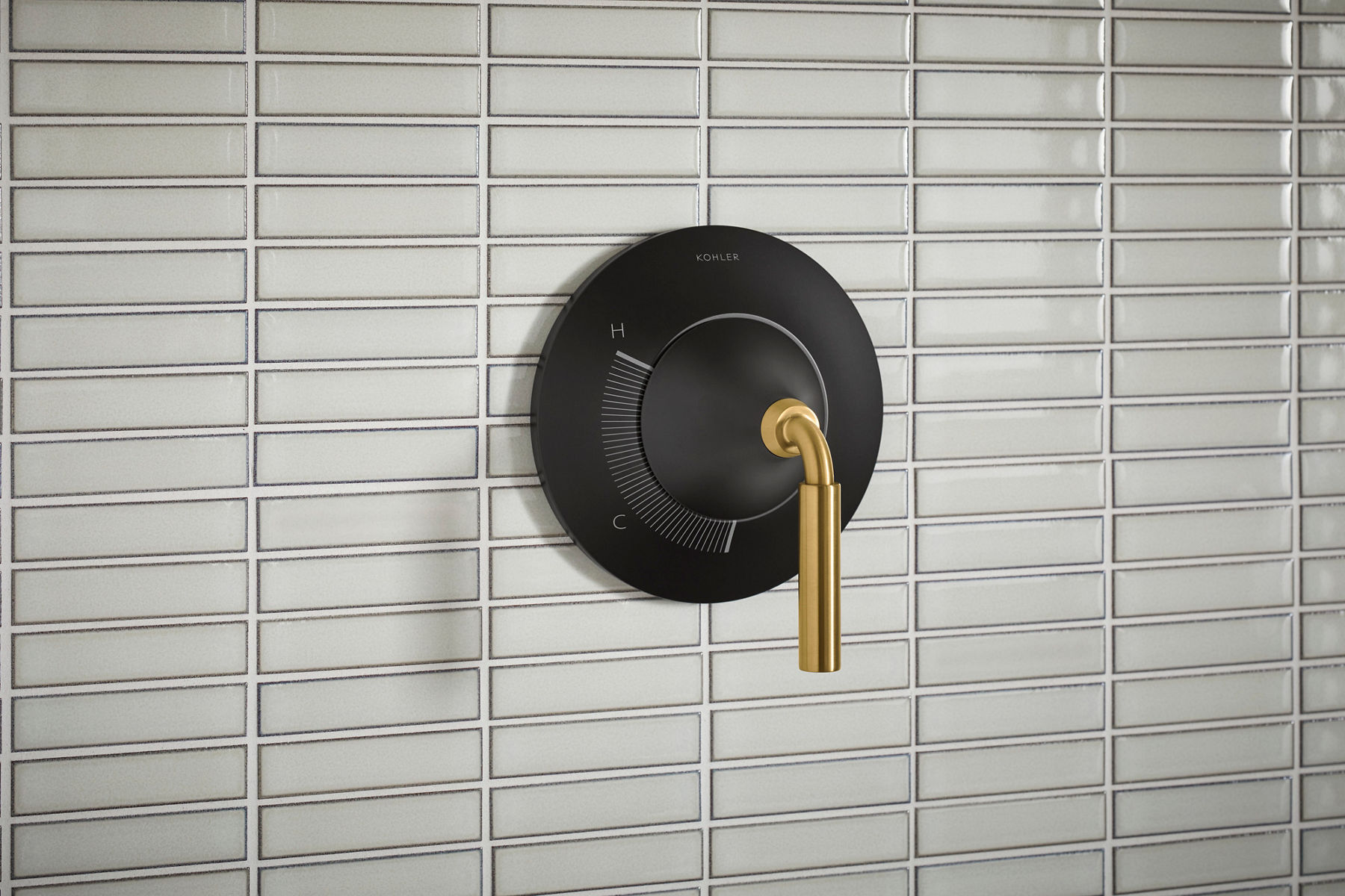 Close-up image of a Kohler Occasion shower control valve handle in two-toned finish featuring matte black wall mount and vibrant brass control handle installed against a small patterned white tile. 