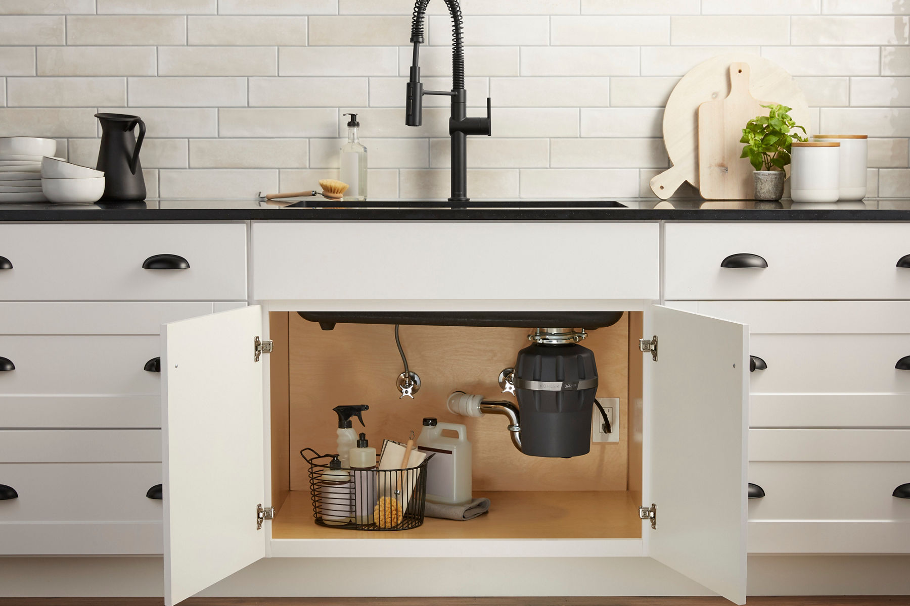 White Shaker style cabinet doors stand open, revealing a KOHLER garbage disposal and a basket of cleaning supplies.