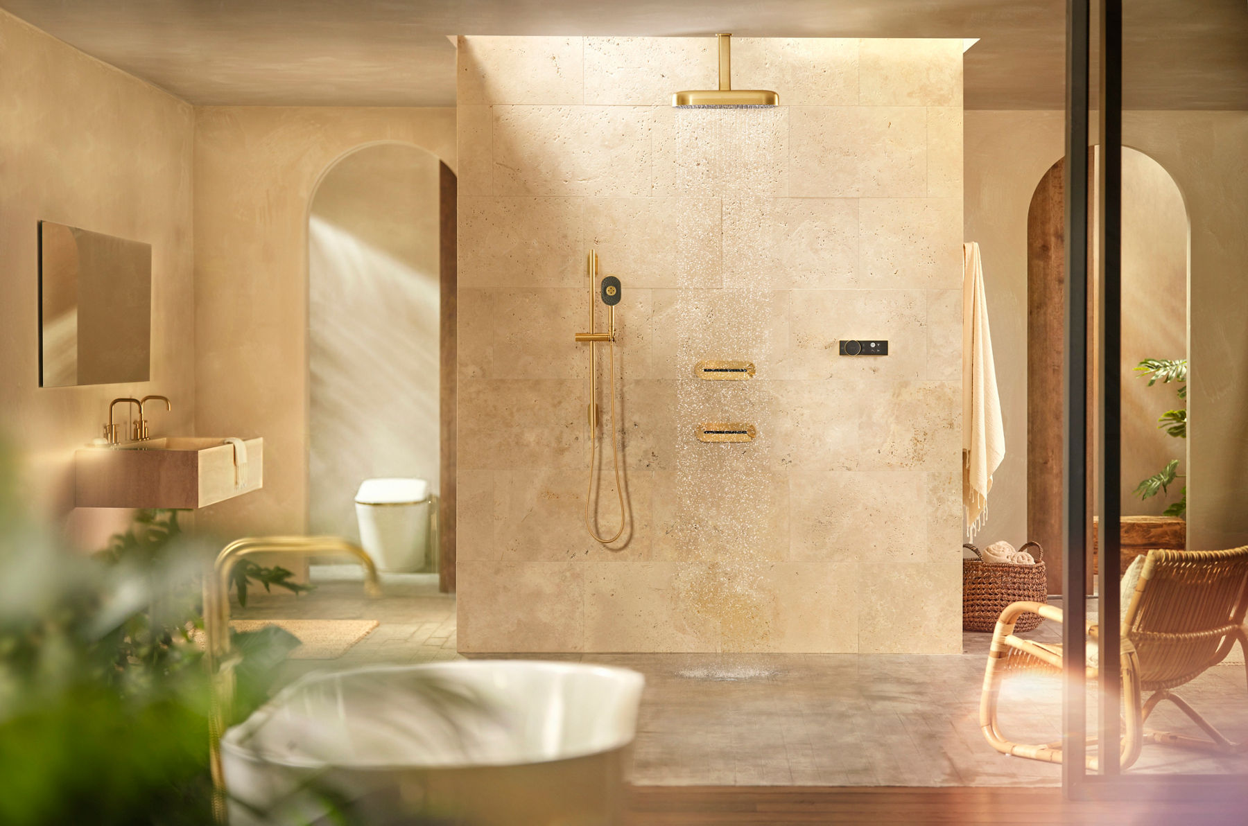 A luxurious shower area with beige tiled walls featuring a gold rainhead shower, handheld shower, and modern fixtures with a partially visible bathroom in the background.