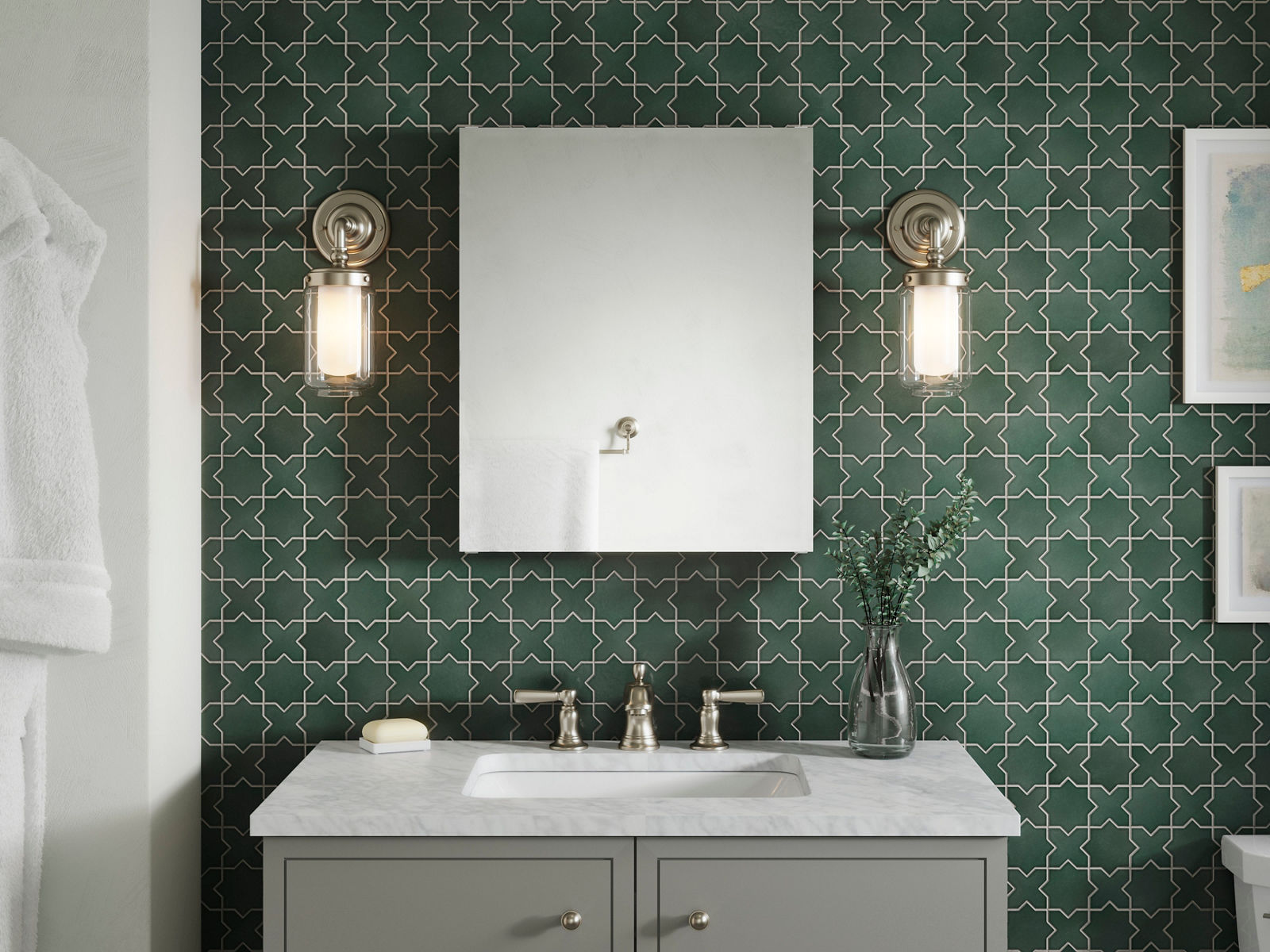 Centered and symmetrical image of a bathroom vanity space with a rectangle medicine cabinet mirror, matching Kohler sconce lighting on both sides of the mirror, and a grey two-door vanity with marble top and polished nickel faucet. A white cloth robe is seen hanging next to the vanity space and a rich green color patterned tile accents the wall.