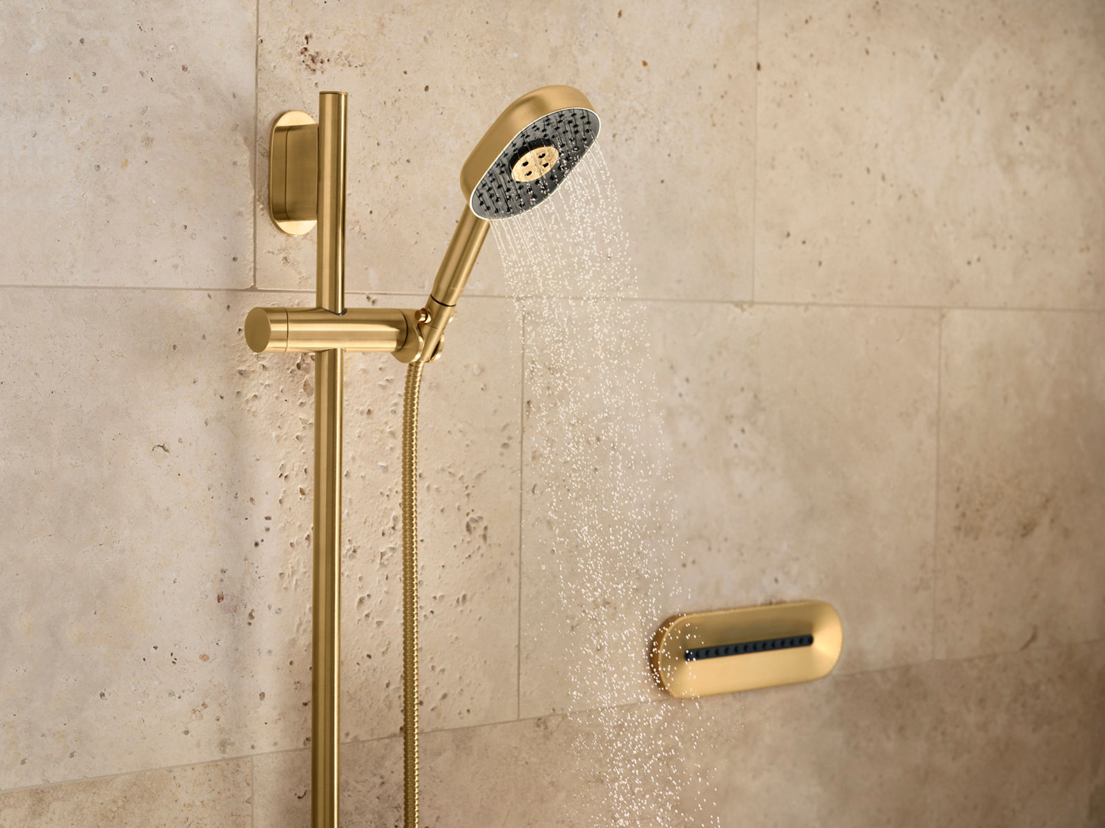 A brushed brass hand shower attached to a sliding rail, mounted to a tan-colored stone wall with an oval-shaped body spray to the side.