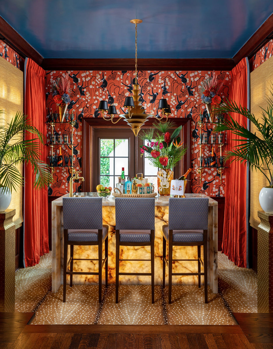 A vibrant home bar with a blacklit onyx countertop, red patterned wallpaper, gold chandelier, and blue upholstered barstools decorated with plants and a variety of bottles.