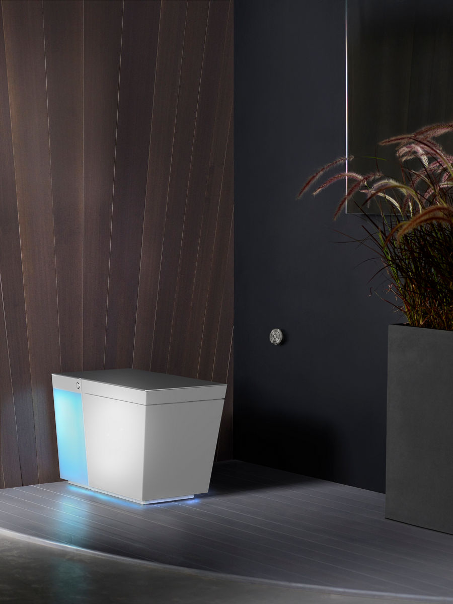 A white Numi toilet with the rear and underside illuminated in a bathroom with one gray wall, one wood-lined room, and gray floors.