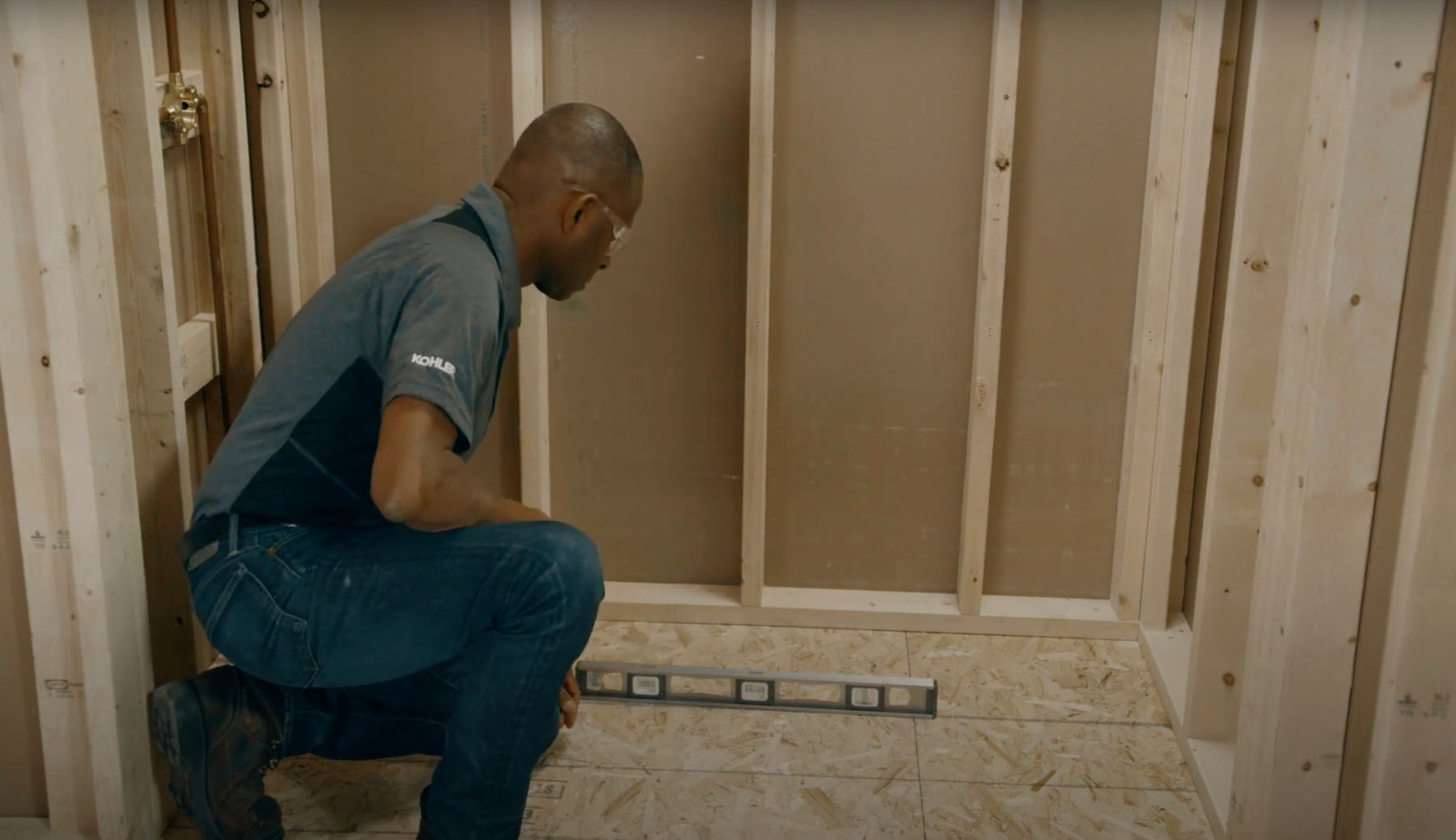 A man in work clothes checking the level of a subfloor in an unfinished bathroom.