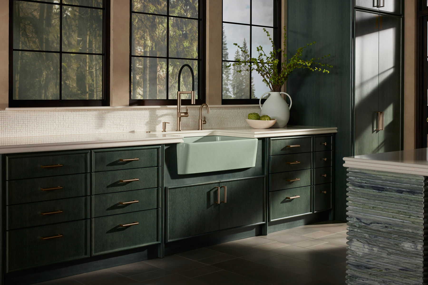 Elegant kitchen with dark green cabinetry, brass hardware, and a large farmhouse sink. A decorative vase with green branches sits on the countertop, and natural light streams in through large windows.