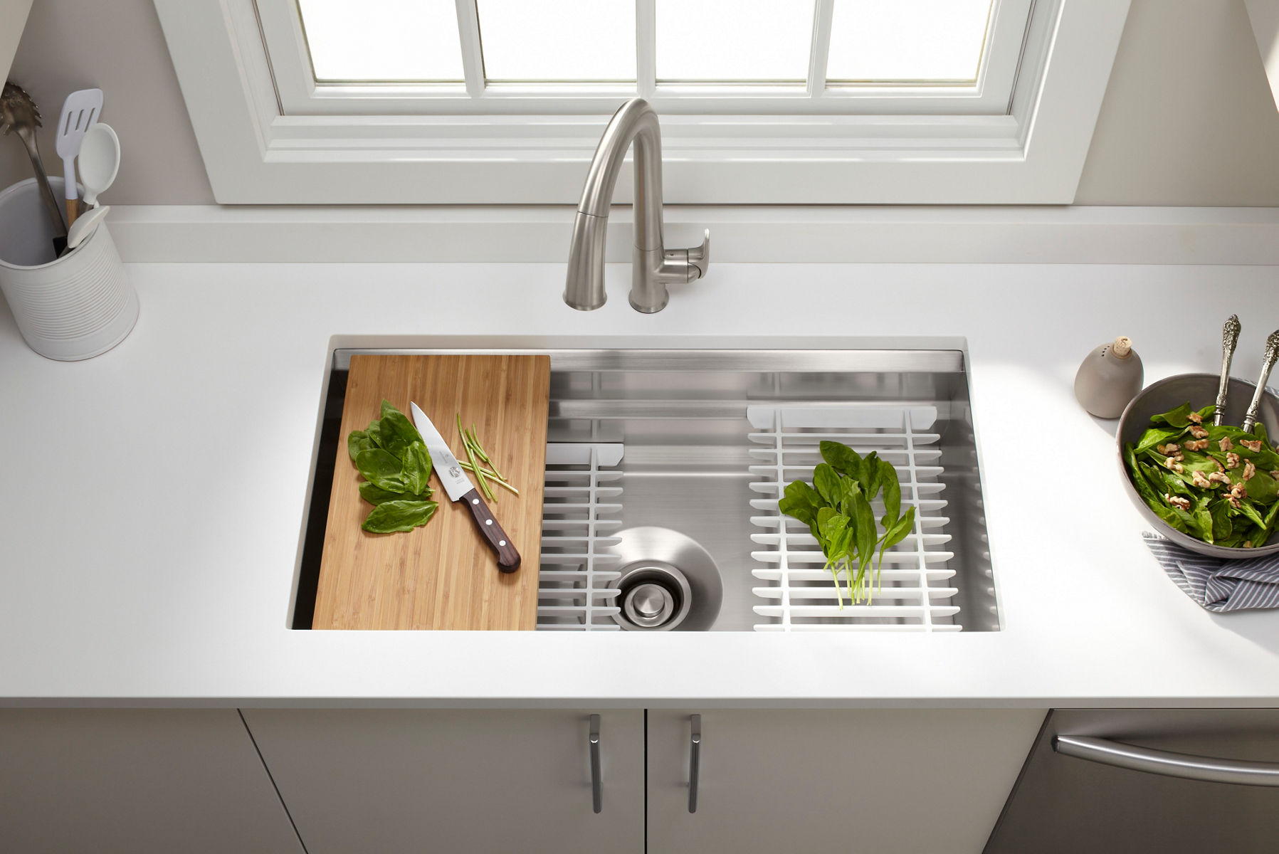A workstation kitchen sink with a cutting board and leafy greens featuring a stainless steel faucet and a window above the sink.