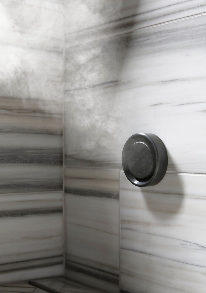 Close up of a steam nozzle emitting steam, mounted to a tiled stone wall with light gray, dark gray, and light brown horizontal streaks.