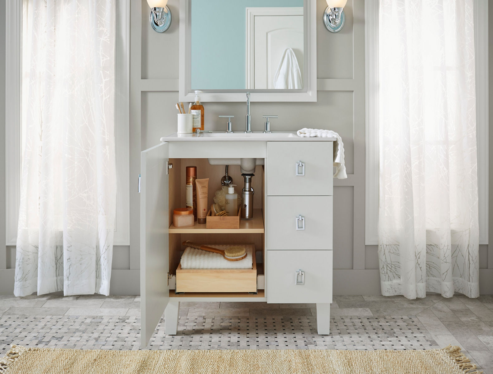 A light gray bathroom vanity against similarly colored coffered wall with floor-length windows and window treatments on either side of the vanity.