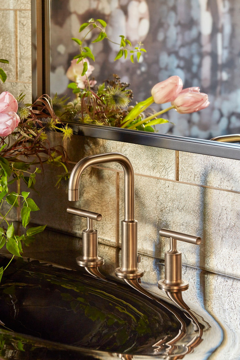 Close up of a widespread faucet in brushed bronze against a stone wall with a mirror, and a flower arrangement on the countertop.