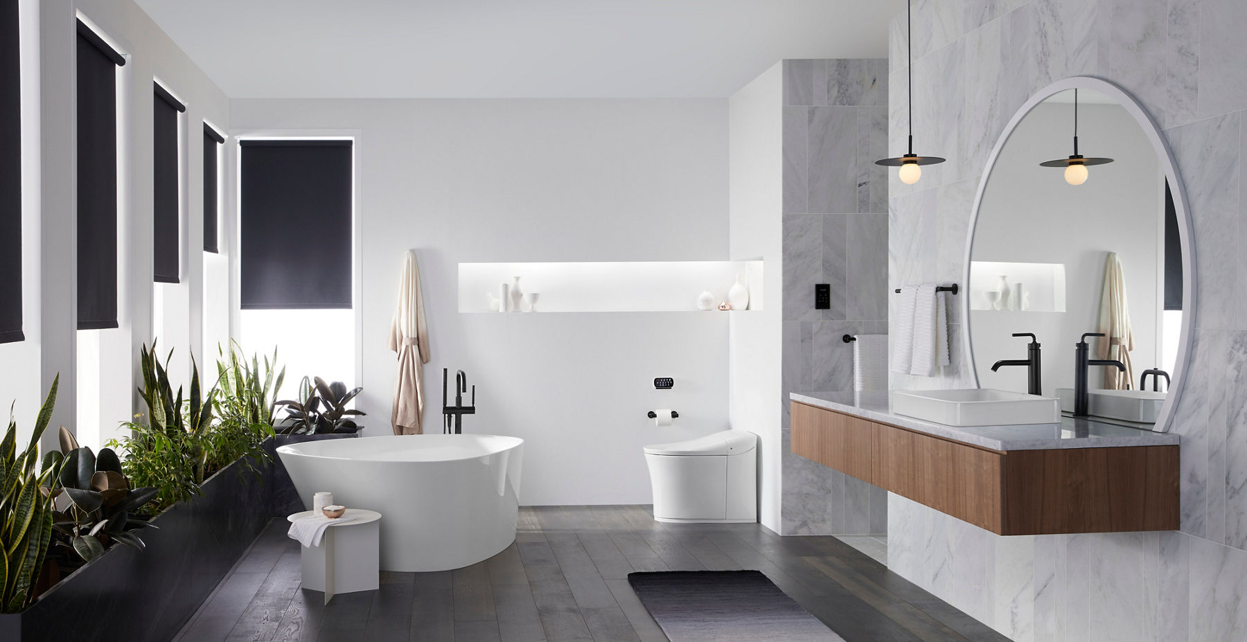 Minimalist bathroom with a freestanding white bathtub, black faucet, modern toilet, and built-in shelf displaying decorative items. The room has dark wood floors, a potted plant, and natural light filtering through a black window shade.