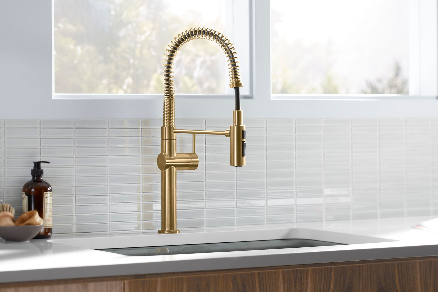 A brushed brass semi-professional kitchen faucet in a white countertop with a white tiled backsplash and a large sun-filled window above.