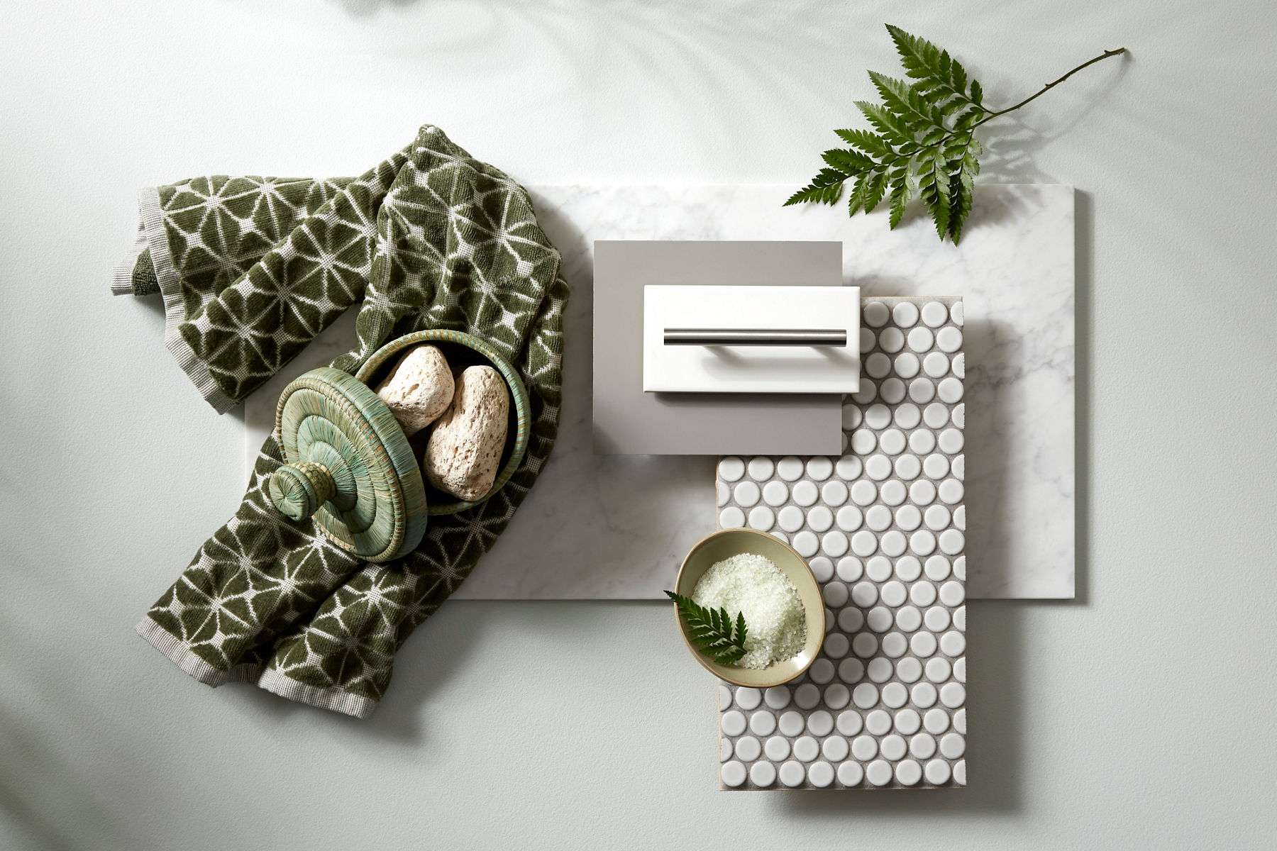 Overhead view of green patterned towel, small green basket, leaf, bowl, cabinet hardware sample, white tile sample, and paint sample on a neutral backdrop.